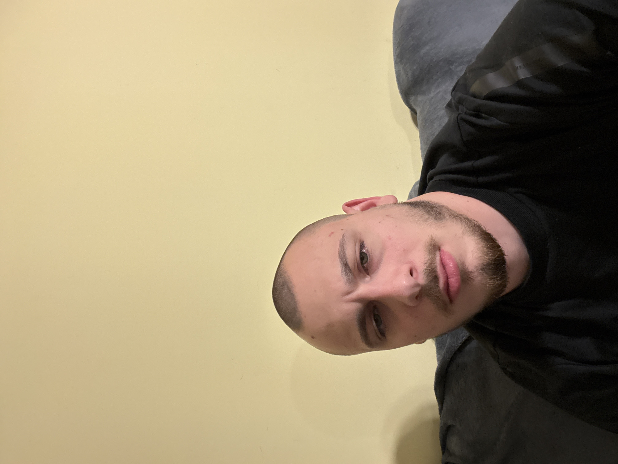 A medium-close shot of a man’s face, taken from a slightly downward angle. The man has a closely shaved head and a short beard. He is fair-skinned. The background is a plain, light yellow wall and a gray garment that is not specified. The location, Stara Zagora, Bulgaria, cannot be verified from the visual information provided.