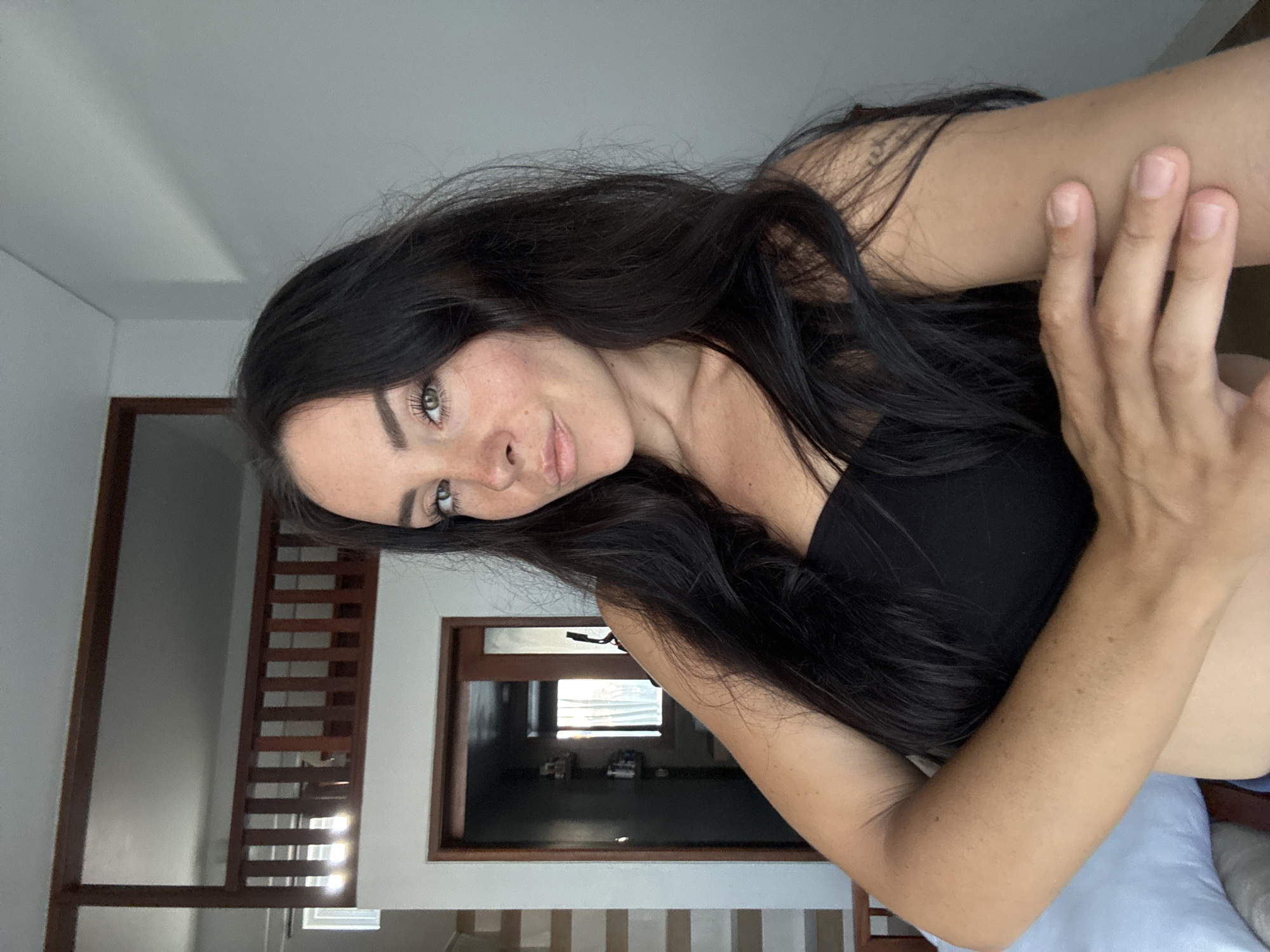 A medium shot shows a woman with long, dark hair in a strapless black top posing for a selfie in what appears to be a bedroom with a loft area. The woman is positioned in the foreground, facing the camera with her left arm resting across her chest and her right arm bent, hand resting on her upper arm. She has fair skin with freckles on her nose and cheeks, and her eyes are green. Behind her, a wooden railing and stairs suggest a loft or second floor. To her left, a doorway leads into another room. The walls are white, and natural light streams in from a window on the far left, casting shadows on the wall. The overall impression is of a casual, indoor setting, likely a residential space in North Kuta, Indonesia, given the architectural style and ambiance.