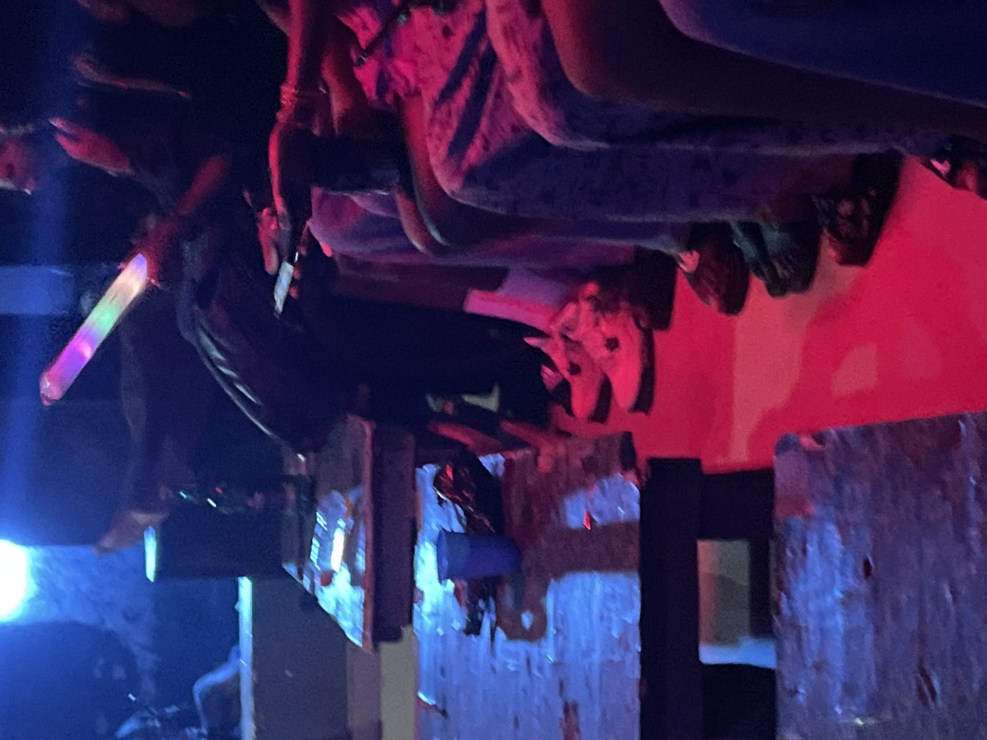 In Durumi, Nigeria, a group of individuals are gathered in a dimly lit indoor setting, possibly a club or a lounge. The environment is characterized by vibrant, atmospheric lighting, with prominent blue and red hues illuminating the scene. Several people are seated at tables, with some holding electronic devices, suggesting they are engaged with their phones. One individual is holding a glow stick that is emitting multi-colored light. The tables themselves appear to be made of dark wood and are cluttered with various objects, including a blue plastic cup and what seem to be wrappers or discarded items. The overall impression is of a social gathering or entertainment event taking place in a low-light, lively environment.