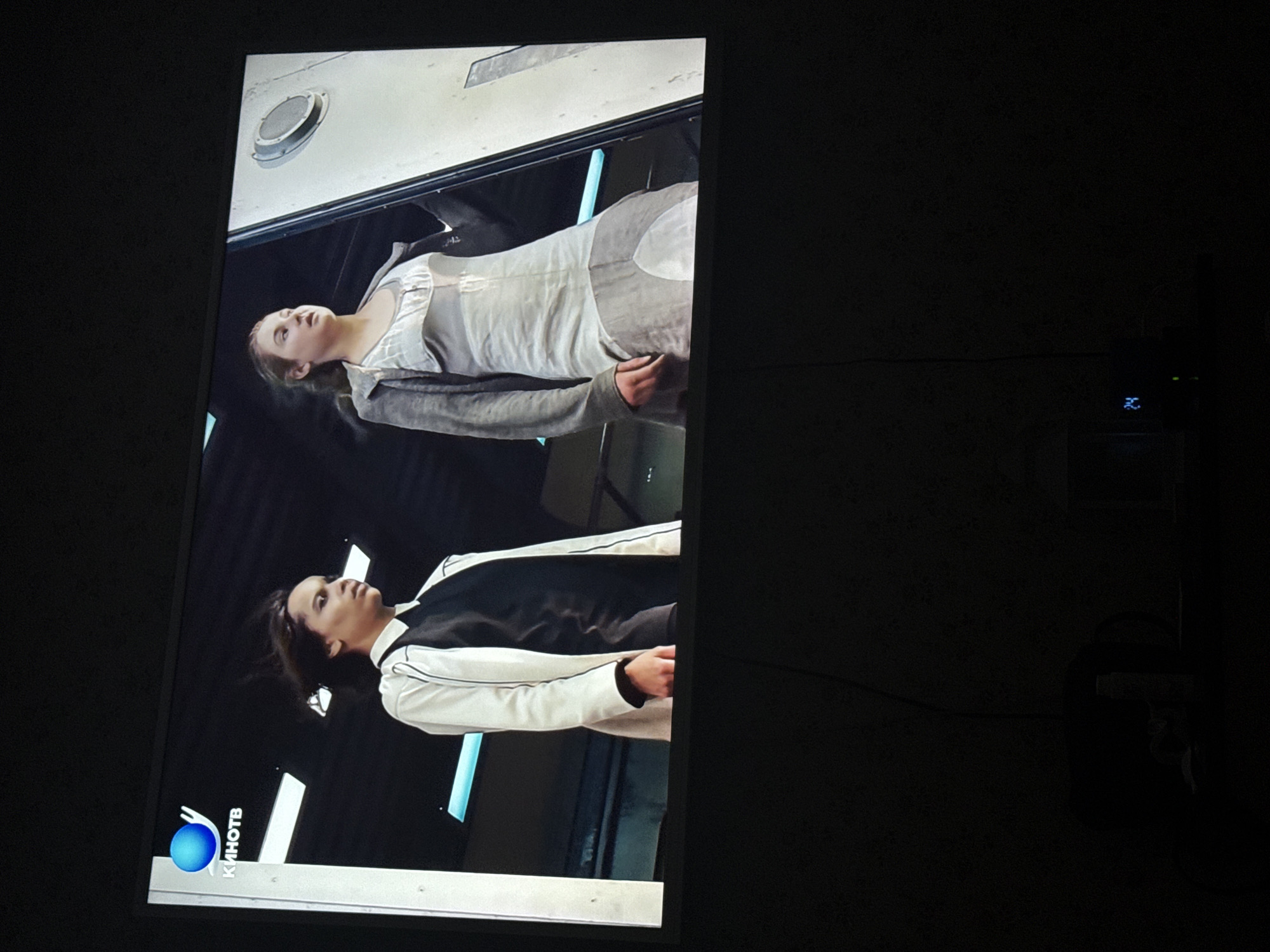 The image shows a television screen displaying a scene with two women. The woman at the bottom has dark hair and is wearing a black shirt with a white jacket. The woman at the top has fair hair and is wearing a light-colored, layered outfit. The television is broadcasting content with the logo of a Russian television channel, "KINOTV," visible in the lower-left corner. The television itself is in a dark room. Although the channel is available in Mytishchi, Russia, the location of the photo is not directly identifiable as Mytishchi.