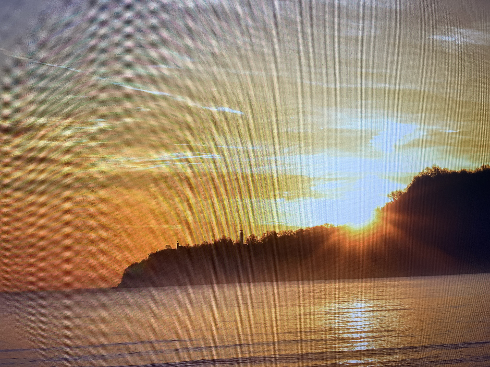This image depicts a serene coastal landscape during either sunrise or sunset, likely near Varna, Bulgaria. The sun, a brilliant source of light, is positioned behind a dark, forested cliff or hillside, creating a dramatic sunburst effect and casting warm, golden rays across the scene. The sky is painted with vibrant hues of orange and yellow near the horizon, fading into softer blues and grays higher up, with delicate clouds streaking across.
The silhouette of the forested land stretches across the middle ground. On its crest, two distinct structures are visible: one on the left appears to be a lighthouse, while another, taller tower stands further to the right. Both are starkly silhouetted against the bright sky. The foreground is dominated by a calm body of water, likely the Black Sea, which beautifully reflects the warm colors of the sky and the sun's light, showing gentle ripples and shimmering reflections.
The overall atmosphere is peaceful and majestic, characteristic of dawn or dusk. A notable detail of the media file itself is the visible pixel grid pattern, indicating that the image is a photograph of a digital screen rather than a direct capture of the scene. There is no visible text in the image.