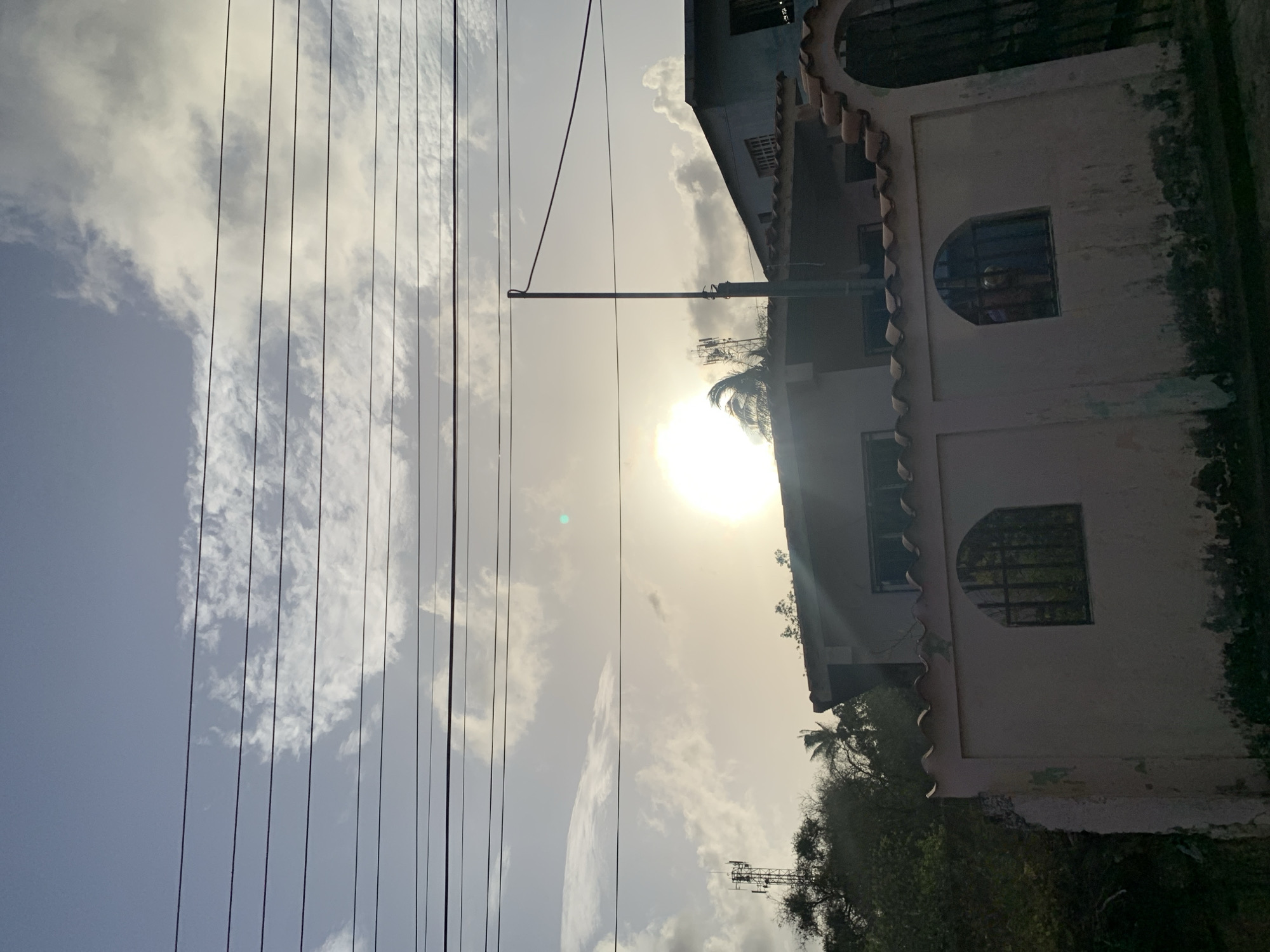 The image captures a street scene at dusk in Santa Ana, Venezuela. In the foreground, a light pink, weathered wall runs horizontally, featuring two arched windows with dark metal bars. Above the wall, power lines are strung across the frame, silhouetted against a pale blue sky filled with scattered white clouds. The sun, a bright, hazy orb, is visible in the upper middle portion of the image, casting a warm glow. Behind the wall and partially obscured by trees and foliage, a two-story building with a tiled roof is visible, along with a dark metal gate. In the distance, a cell tower stands tall, and other buildings are faintly discernible. The overall impression is one of a residential area during twilight.