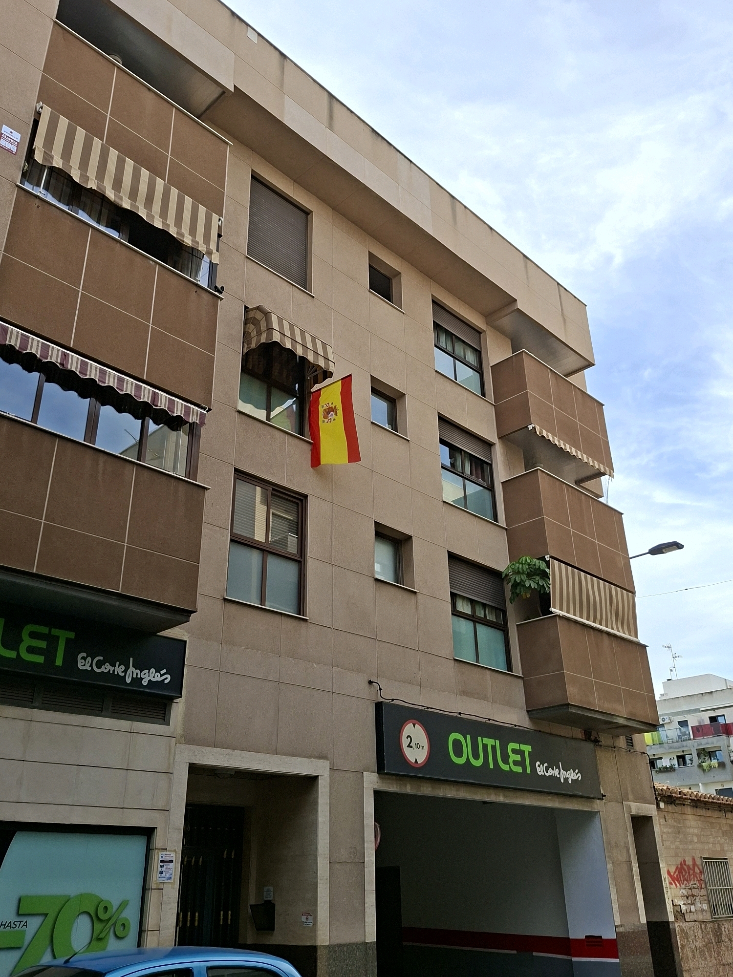 The image shows the facade of a multi-story building in Torrevieja, Spain. The building has a beige, tiled exterior and features several balconies with brown awnings. A Spanish flag is displayed from one of the windows. Below the residential levels, there is a commercial space with signage for "OUTLET El Corte Inglés." A blue car is partially visible in the foreground. The sky is cloudy.