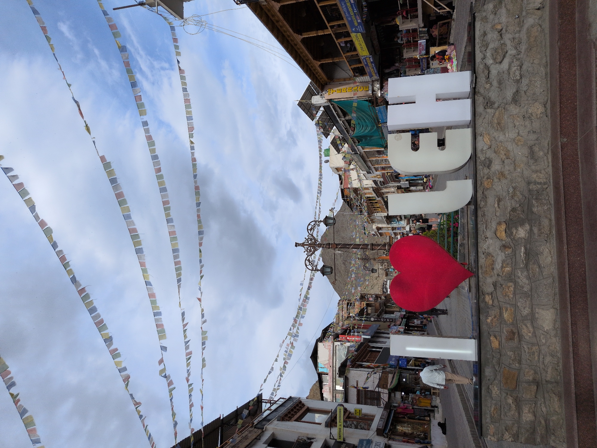 A street scene in Leh, India, features a large, red heart sculpture and white block letters spelling "LEH." The letters and heart are placed on a stone platform in the foreground. Behind the sculpture, a street is lined with multi-story buildings housing shops and businesses. Prayer flags are strung across the sky, and mountains are visible in the distance. Several people are walking along the street. The sky is cloudy with patches of blue.