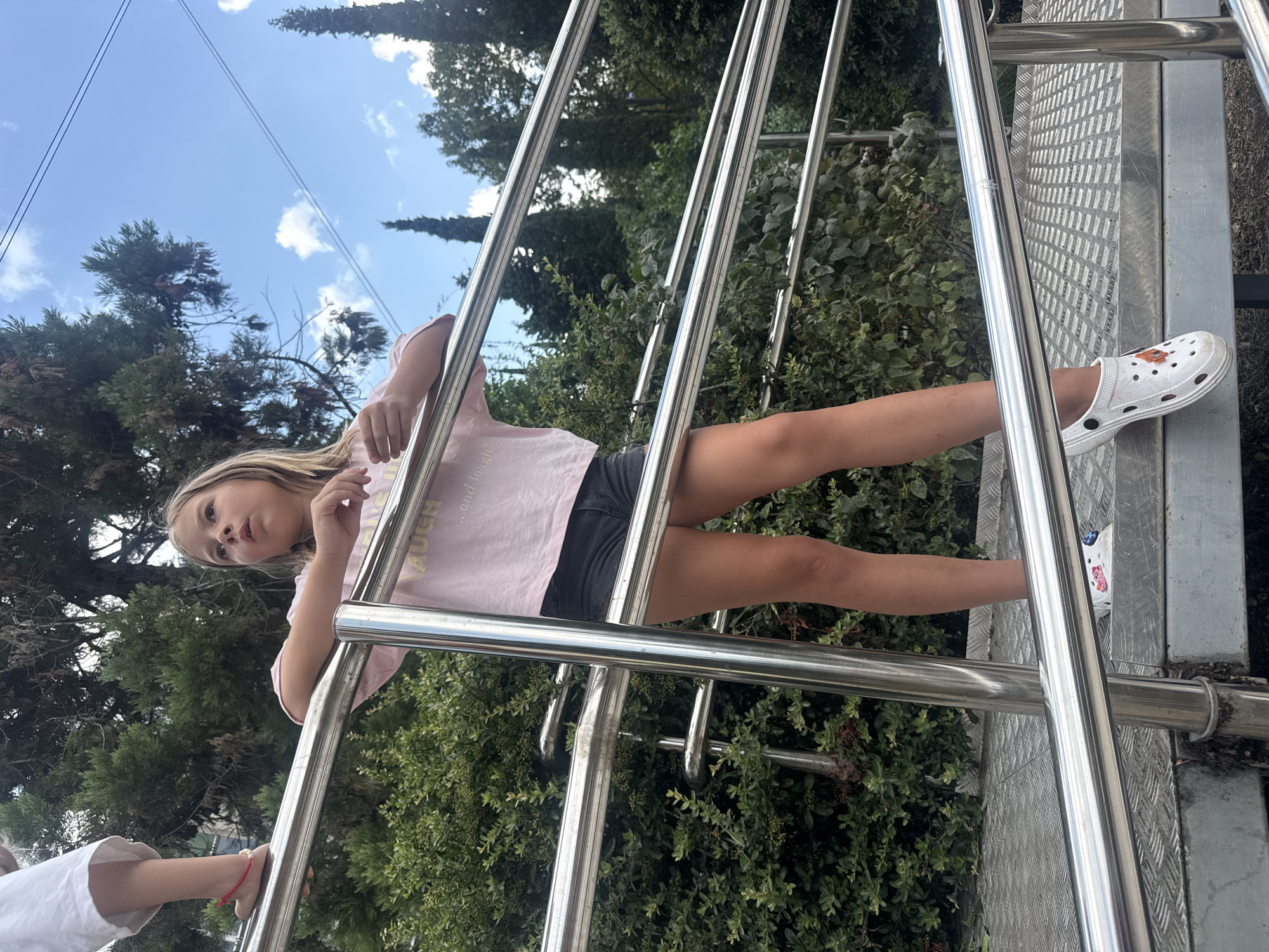 A young blonde girl with a light pink t-shirt and black shorts leans against a metal railing, looking towards the camera. She has her left arm resting on the railing and her right arm is bent with her hand near her face. Her feet are visible, with one wearing a white Croc with an orange design and the other bare. Another person's legs and feet, wearing a white shirt, are visible in the lower left corner. The scene is outdoors, with lush green foliage in the background and a blue sky with scattered clouds visible above. The metal railing appears to be part of a staircase or platform. The girl's expression is somewhat serious or contemplative. The text on her t-shirt is partially visible and appears to say "laugh" and "...and love". The overall impression is a casual, daytime outdoor scene, possibly in a park or garden setting.
