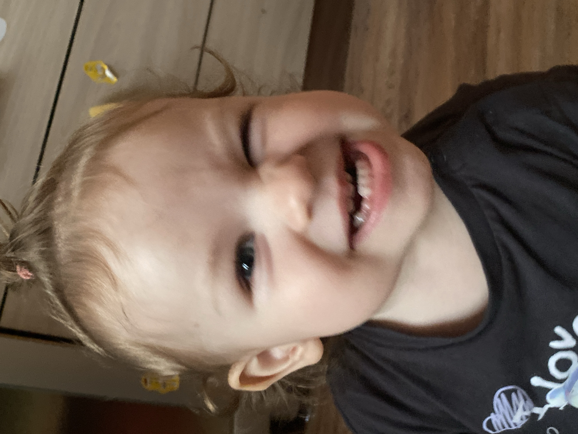 The image captures a close-up, slightly tilted view of a young child, likely a toddler, smiling widely. The child has light-colored hair, possibly blonde or light brown, pulled back with a pink or red hair tie visible on the left side of their head. Their left eye appears partially closed, possibly in a playful wink or squint, while their right eye is open and looking towards the viewer. The child's mouth is open in a broad smile, revealing several teeth.
The child is wearing a dark-colored, short-sleeved t-shirt, which appears to be black or dark grey. Part of a design or text is visible on the shirt, showing white lettering that spells "lov" with what looks like a doodle of a heart next to it, suggesting the word "love".
The background appears to be an indoor setting with a wooden floor or a surface with a wood grain pattern. Some thin dark lines suggest floorboards or seams. Scattered near the child's head are a couple of small, yellow, crinkled objects, possibly candy wrappers or small pieces of paper. The lighting is soft and even, suggesting an indoor environment without harsh shadows. The overall emotion conveyed by the child's expression is one of joy, happiness, or playfulness.