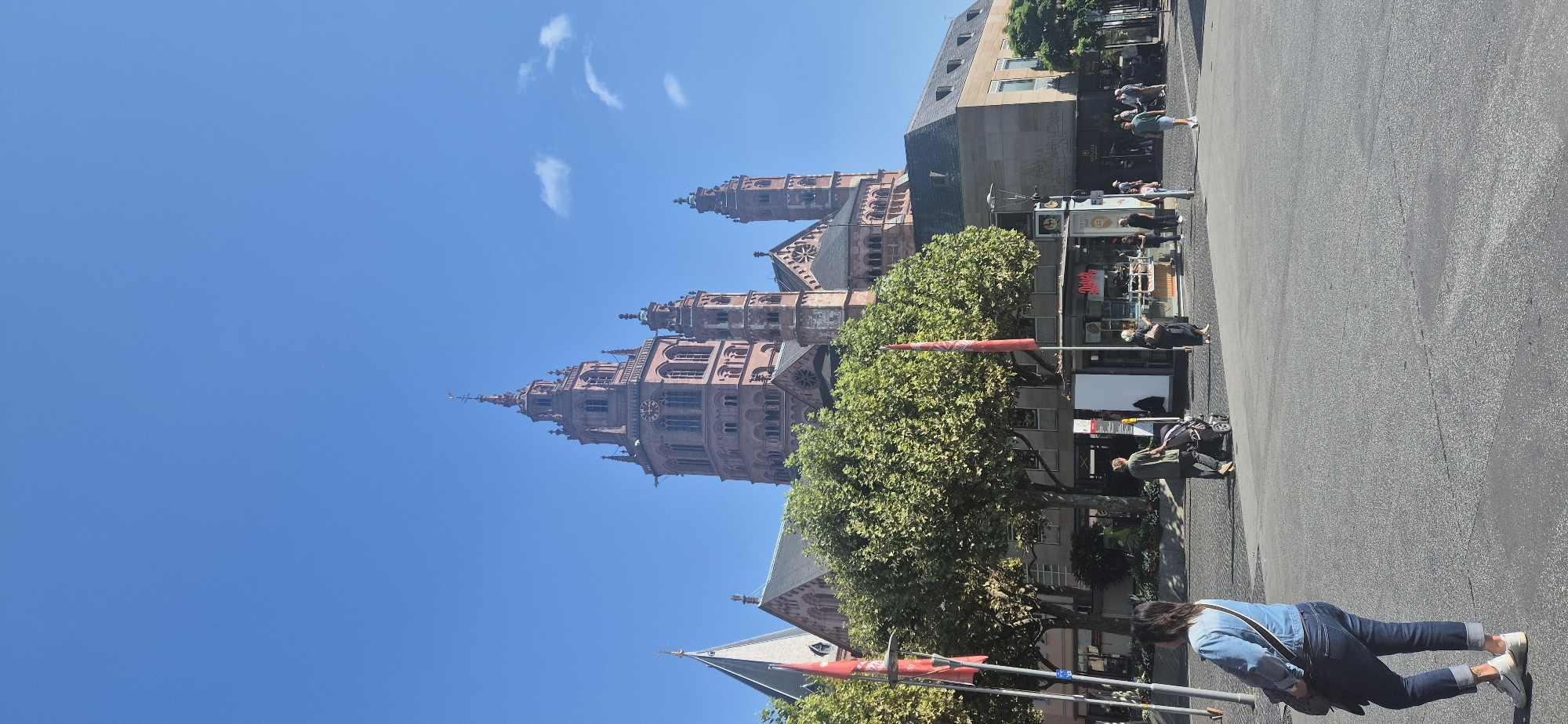 In Mainz, Germany, a wide view captures a bright, clear day with a deep blue sky. Dominating the background is the imposing, reddish-brown stone facade of Mainz Cathedral, its spires and architectural details clearly visible against the sky. In front of the cathedral, a row of lush green trees provides a vibrant contrast. At the base of the trees and extending towards the foreground, a street scene unfolds. Several people are present: a woman in a denim jacket and jeans walks away from the camera, her back to the viewer, carrying a bag over her shoulder. Further in the middle ground, a person pushes a stroller, while other individuals are seen near a kiosk or small shop with colorful signage. A street lamp and red flags on poles are also visible. The ground is paved with asphalt.