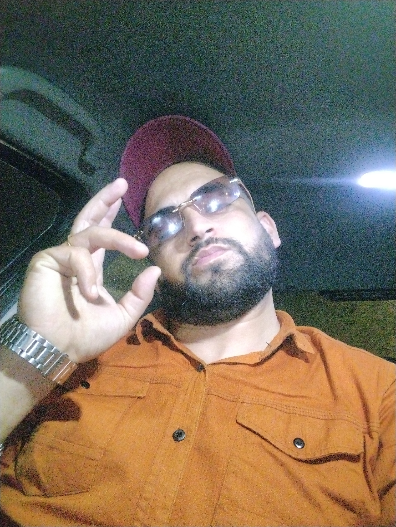 The image features a man wearing a maroon baseball cap, sunglasses, and an orange button-down shirt with two chest pockets. He has a beard and is wearing a watch. He is holding his hand up, with his thumb and index finger close together. The background is dark, suggesting an indoor environment, possibly a car. The location, Martil, Morocco, cannot be determined from the image alone.