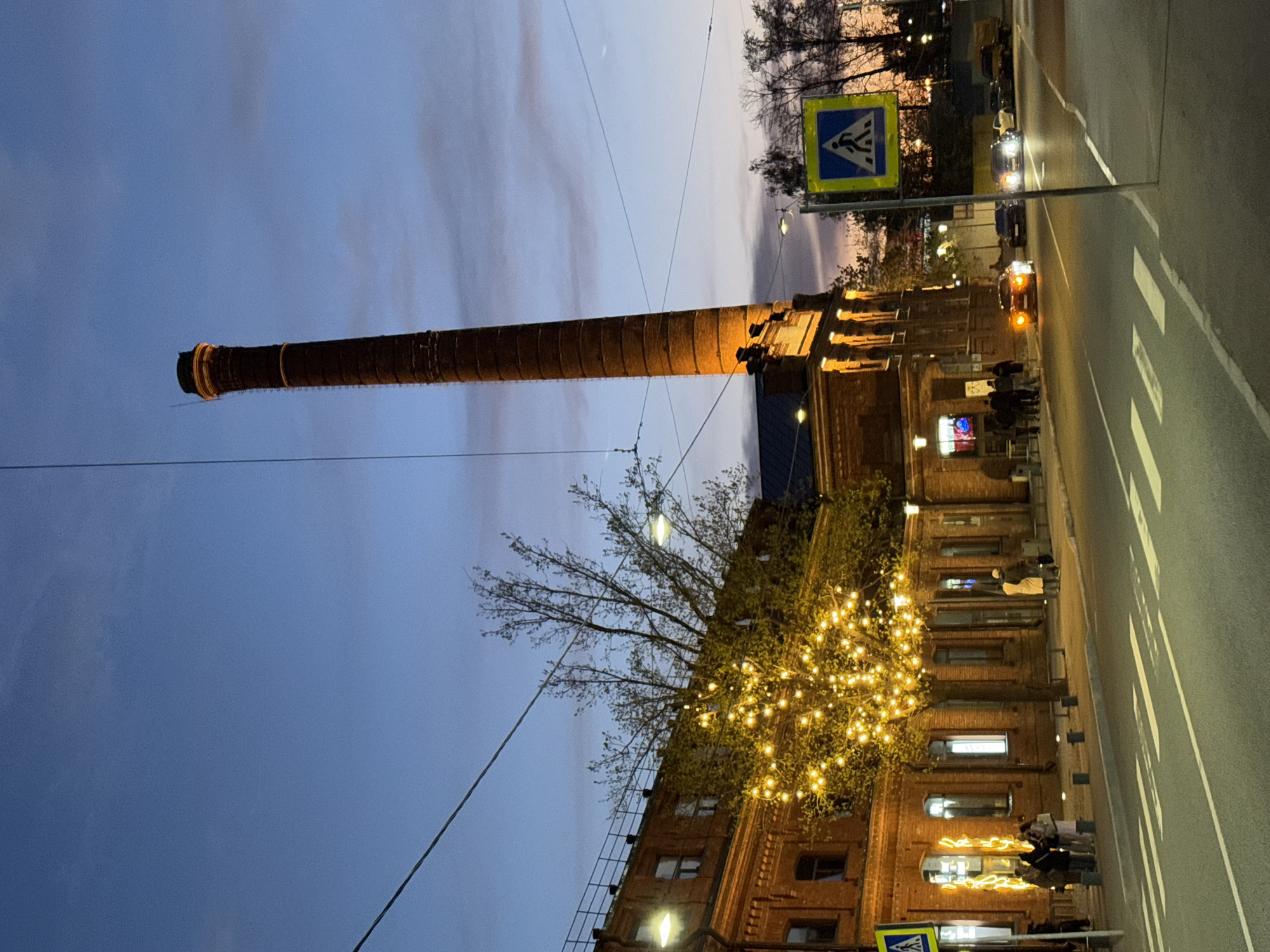 The image depicts a nighttime urban scene in Saint Petersburg, Russia. A tall brick chimney dominates the left side of the frame, likely part of an old industrial complex. Adjacent to the chimney is a brick building illuminated by warm lights, with trees adorned with decorative lights in the foreground. A street runs along the right side, with a crosswalk and pedestrian sign visible. Cars with headlights on are visible on the street. People are walking or standing near the building. The sky transitions from blue to a light orange-pink at the horizon, indicating either dusk or dawn.