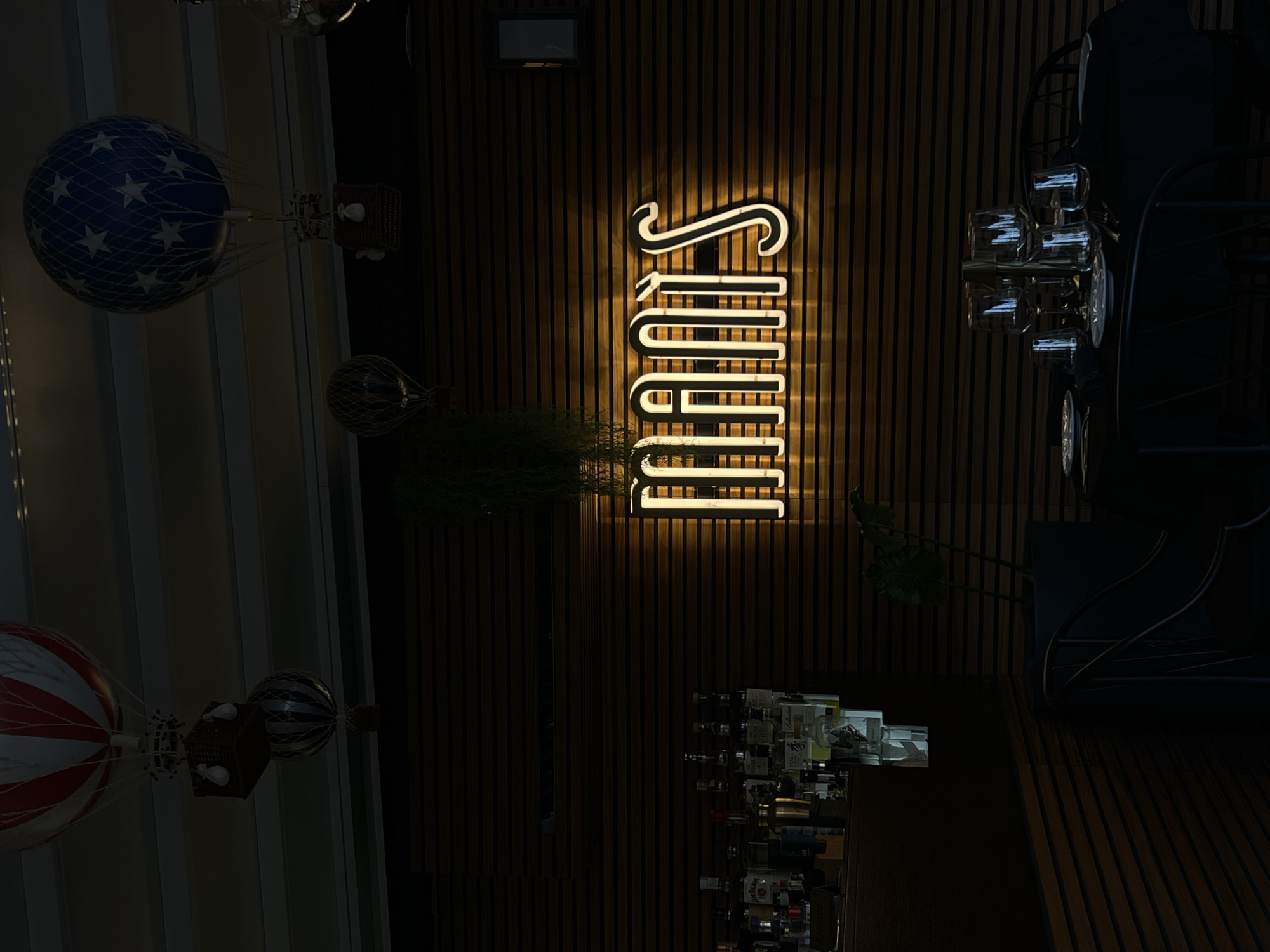 The image depicts an interior scene of a restaurant or bar with a dimly lit ambiance. The background features a wall with horizontal wooden paneling. Suspended from the ceiling are several decorative hot air balloon models, one with red and white stripes, another blue with white stars, and two smaller, darker ones.
Prominently displayed on the wooden wall is a bright, neon sign that spells out "MANIS" in a stylized, white, outlined font with a yellow glow. To the left of the sign, a bar area is visible, stocked with various bottles of alcohol and glasses. In the foreground, a dark-covered table is set for dining with plates, glassware, and chairs. A potted plant with large leaves is situated to the right of the table. The overall impression is one of a sophisticated and atmospheric establishment. There are no people visible in the image. The location is not specified as Rome, Italy, or any other city.