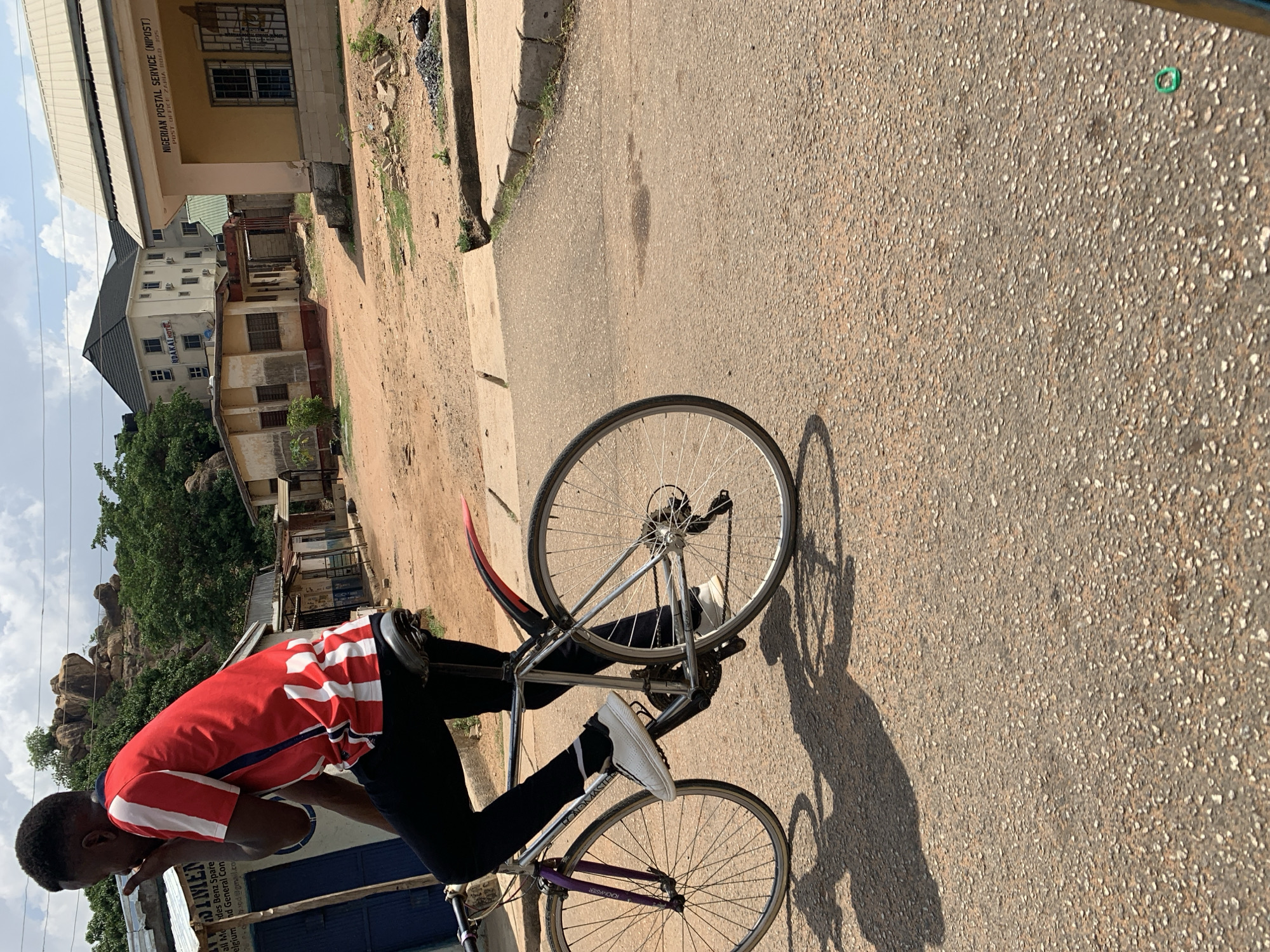 The image depicts a person standing over a bicycle on a paved road. The person is wearing a red and white striped shirt, black pants, and white shoes. The bicycle is silver with purple accents. The background includes buildings and a rocky hill. Signage indicates the presence of the "NIGERIAN POSTAL SERVICE (NIPOST)" and "NDAKAI HOTEL", suggesting the scene is located near these establishments. The image is taken in Rafin Par, Nigeria.