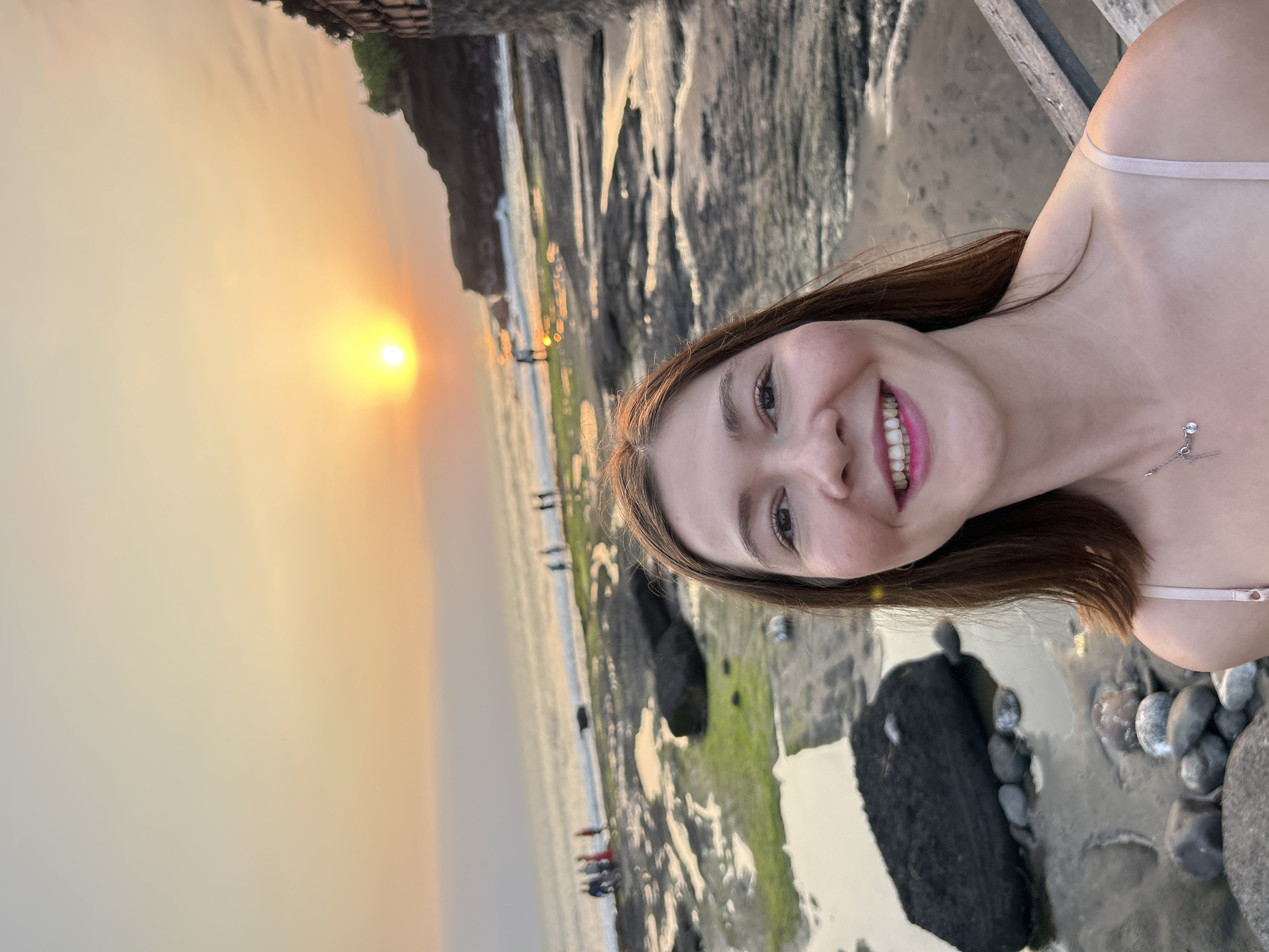 A woman is taking a selfie on a rocky beach at sunset. The sun is low in the sky, casting a warm orange glow over the scene. In the background, the ocean meets the shore, and several people are visible walking along the water's edge. The woman is smiling and looking at the camera. She is wearing a pink slip dress and a silver necklace. The beach is covered in rocks of various sizes, some of which are partially submerged in water. There is also some green algae growing on the rocks. To the right of the frame, a stone structure and some wooden railings are visible. The location is identified as a beach in the city of Kediri, Indonesia.