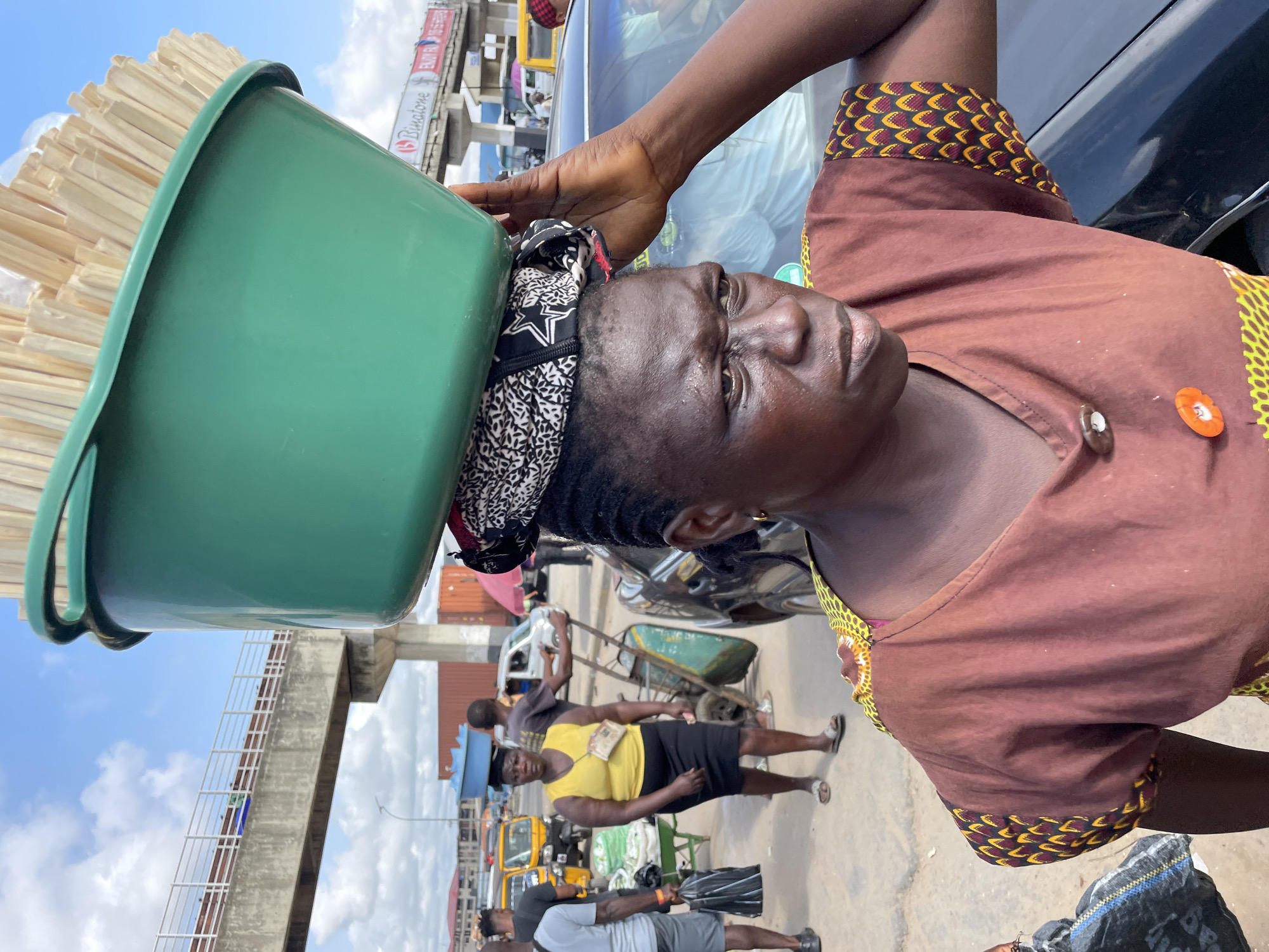 In Onitsha, Nigeria, a woman stands facing forward, her gaze directed slightly to the right, her expression weathered and determined. She is balancing a large, green plastic basin filled with wooden skewers on her head, a traditional method of carrying goods in the bustling city. A patterned headscarf with black, white, and grey designs is positioned beneath the basin, adding a layer of padding. Her attire consists of a brown, short-sleeved dress adorned with a vibrant, patterned fabric along the neckline and cuffs, featuring yellow, red, and black geometric shapes. Two buttons, one brown and the other orange and circular, are visible on the front of her dress.
The background reveals a busy street scene typical of Onitsha. To her left, another woman in a yellow top and black skirt walks with a blue basin on her head, while behind her, a man pushes a green wheelbarrow. Yellow mini-buses, known as "keke napep," and other vehicles populate the street, suggesting a lively commercial area. An elevated pedestrian bridge looms overhead, a modern architectural element contrasting with the traditional scene below. Containers and buildings are visible in the distance, indicating the urban environment. The sky is partly cloudy, with a bright, hazy sun casting a warm light on the scene.