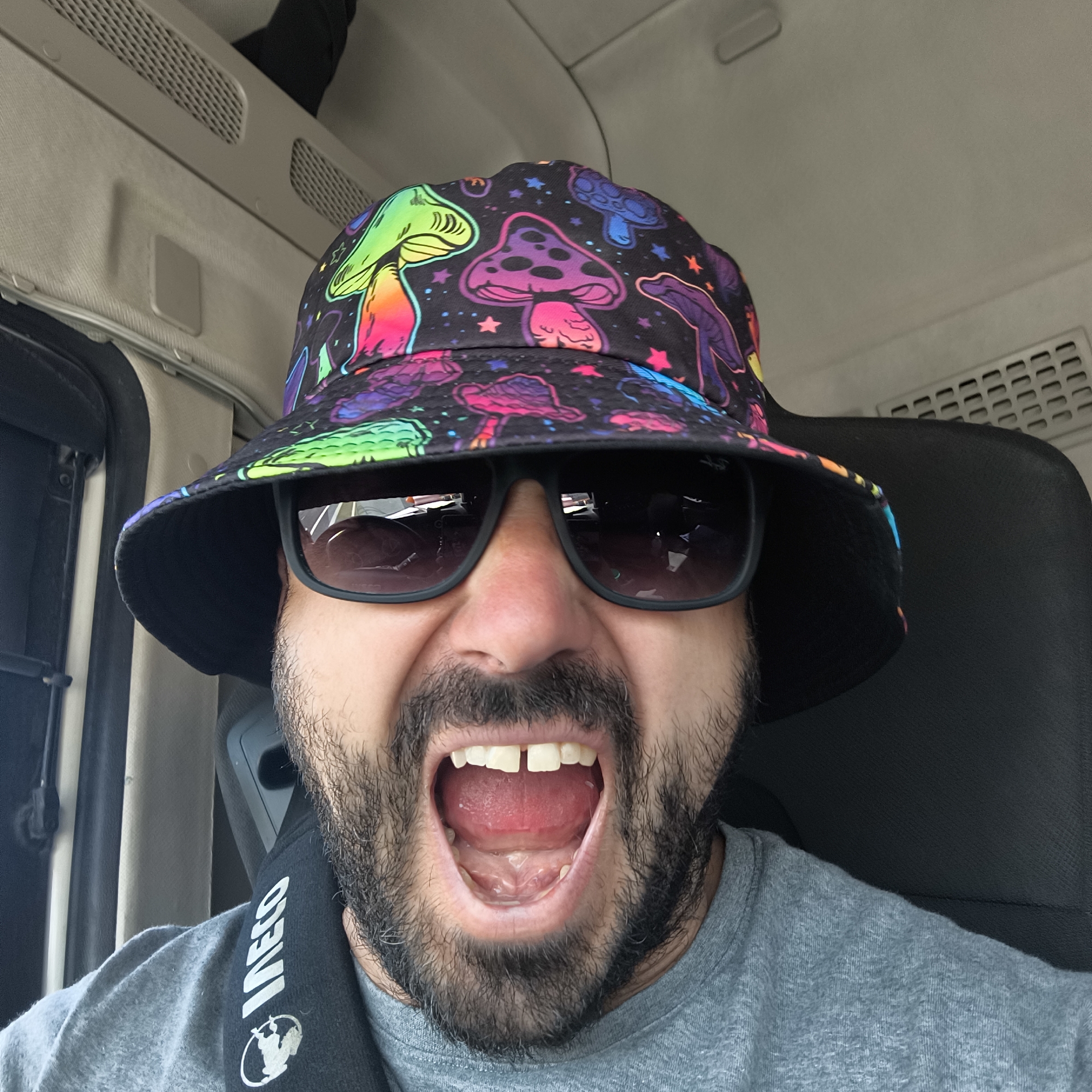 A close-up selfie shows a bearded man with an energetic, wide-open mouth expression inside a vehicle, likely a truck or large van, in Manises, Spain. He is wearing a black bucket hat adorned with vibrant, multicolored psychedelic mushroom patterns and small stars, and dark sunglasses with rectangular frames. His mouth is open wide, revealing his upper and lower teeth, with a distinct gap between his two front upper teeth.
He wears a grey t-shirt, and a black seatbelt crosses his chest. Visible on the seatbelt in white text is the brand "IVECO" and a logo underneath it, resembling a stylized human figure or emblem. The interior of the vehicle, visible behind and above him, consists of a light grey or beige ceiling with ventilation grates. A dark seat with a headrest is also partially visible behind him. The lighting suggests it is daytime. The overall impression is one of excitement or playfulness during travel.