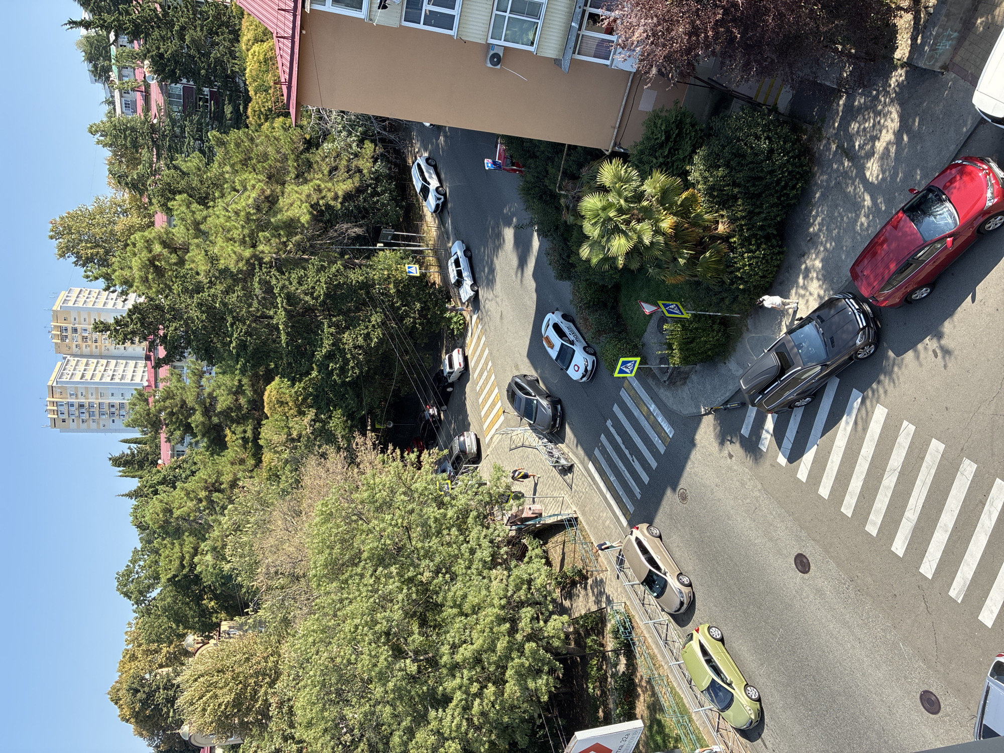 This is an aerial, eye-level, wide-angle shot of a street intersection in Sochi, Russia, on a clear, sunny day. The scene captures a street with multiple cars, pedestrians, crosswalks, and lush green trees. In the foreground, a red SUV is parked on the right side of the road, and a dark SUV with a roof rack is parked behind it, facing away from the viewer. Further up the road, there are several other vehicles, including a white SUV, a white sedan, and a beige hatchback. On the left side of the road, a bright green hatchback is parked. Two pedestrians are visible walking on the sidewalk near a crosswalk. The background features a large building with red tile roofing and tall, slender evergreen trees. In the distance, a tall, multi-story apartment building with many windows is visible. The street is paved and marked with white lines for traffic lanes and crosswalks. The environment appears to be an urban residential area with abundant vegetation.