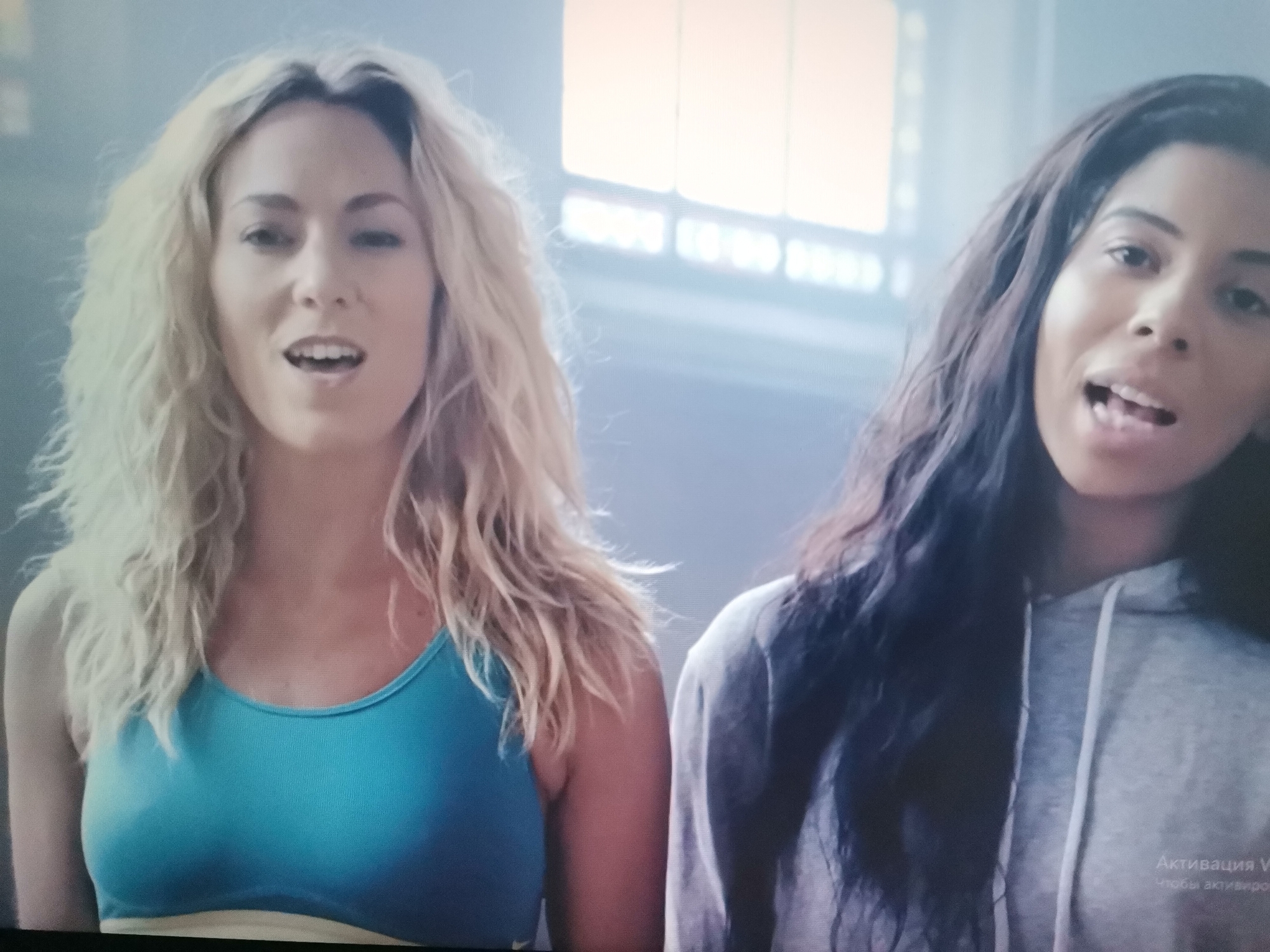 The image shows two women, one with blonde curly hair and the other with dark wavy hair, standing side-by-side in what appears to be an indoor setting with diffused lighting. The woman on the left is wearing a blue sports bra, and the woman on the right is wearing a grey hoodie. Both women have their mouths open, as if speaking or singing. There is text on the lower right of the image that appears to be in Russian, reading "Активация Windows" which translates to "Windows activation". There is no visual information in the image to suggest the location is Belaazyorsk, Belarus.
