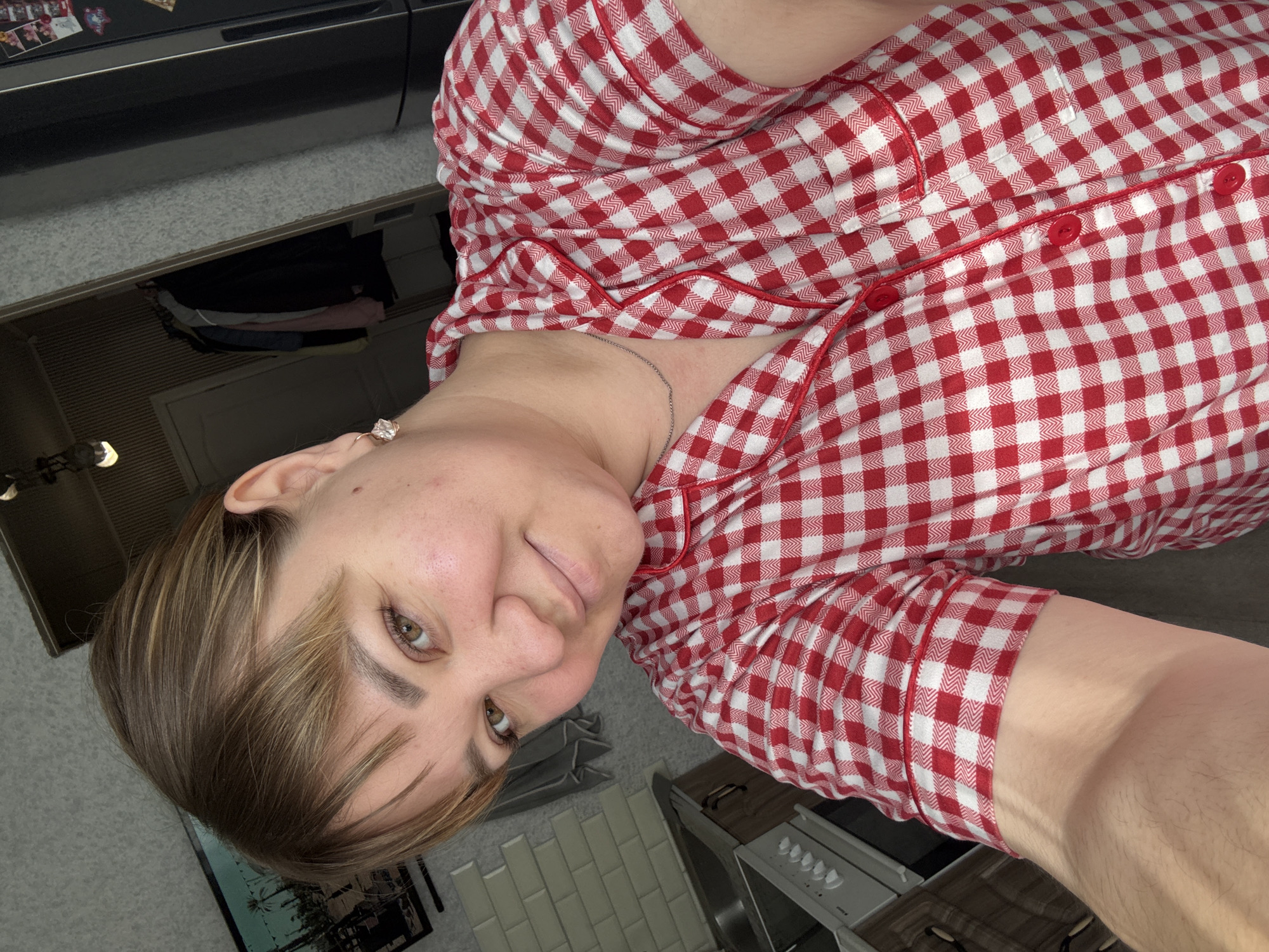 A woman with light brown hair and green eyes is taking a selfie in a bedroom. She is wearing a red and white checkered pajama top. The background shows a kitchen area with a sink and a stove, and a closet with clothes hanging inside. The image was taken in the city of Stary Oskol, Russia.