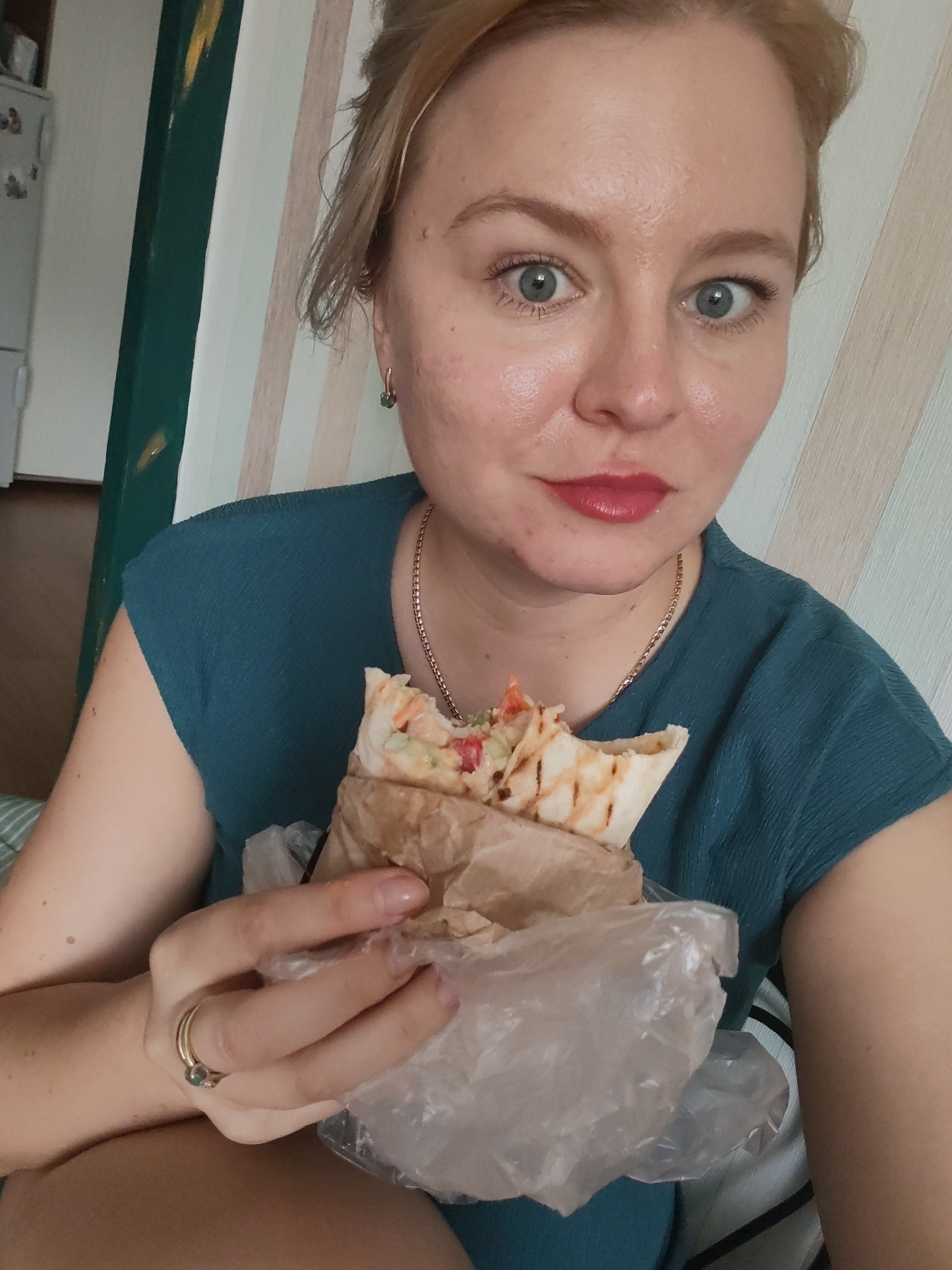 A medium shot photograph, taken indoors, shows a woman holding a partially eaten shawarma. The woman, with blonde hair pulled back and visible ears, has light blue eyes and is wearing a dark teal blouse and a gold necklace. She is also wearing two rings on her left hand, one of which has a small green stone. The shawarma is wrapped in brown paper and then in a clear plastic bag. The background features a wall with vertical cream and light beige stripes, and a dark green wooden beam is visible on the left side of the frame. A white refrigerator is partially visible in the upper left corner. The scene is likely set in an apartment or residential building in Izhevsk, Russia.