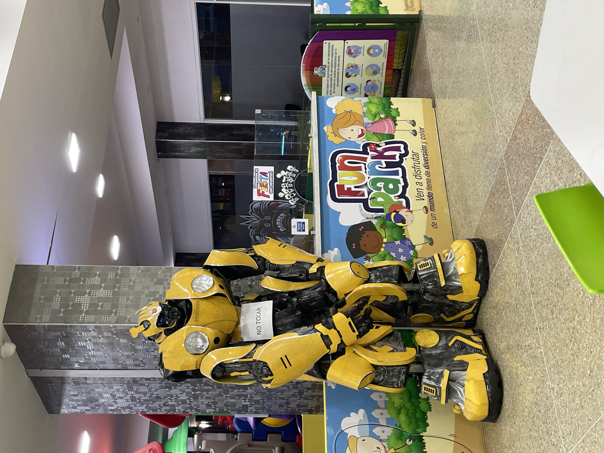 In El Valle del Espíritu Santo, Venezuela, a large, yellow and black robot resembling Bumblebee from the Transformers franchise stands prominently in what appears to be an indoor entertainment venue. The robot is positioned in front of a colorful mural depicting cartoon children and the "Fun Park" logo. A white sign with black lettering reading "NO TOCAR" (Do Not Touch) is attached to the robot's chest. Behind the robot and mural, a glass partition separates this area from another part of the venue, where a sign advertising "Organiza tu FIESTA con nosotros!" (Organize your PARTY with us!) is visible. The environment includes a tiled floor, a large pillar with a patterned texture, and overhead lighting. To the right, a section of a purple display board with circular images and text about children and supervision is partially visible. A white table and a bright green chair are in the foreground, suggesting a seating area. The overall scene depicts a children's play or party venue with a notable robotic mascot.