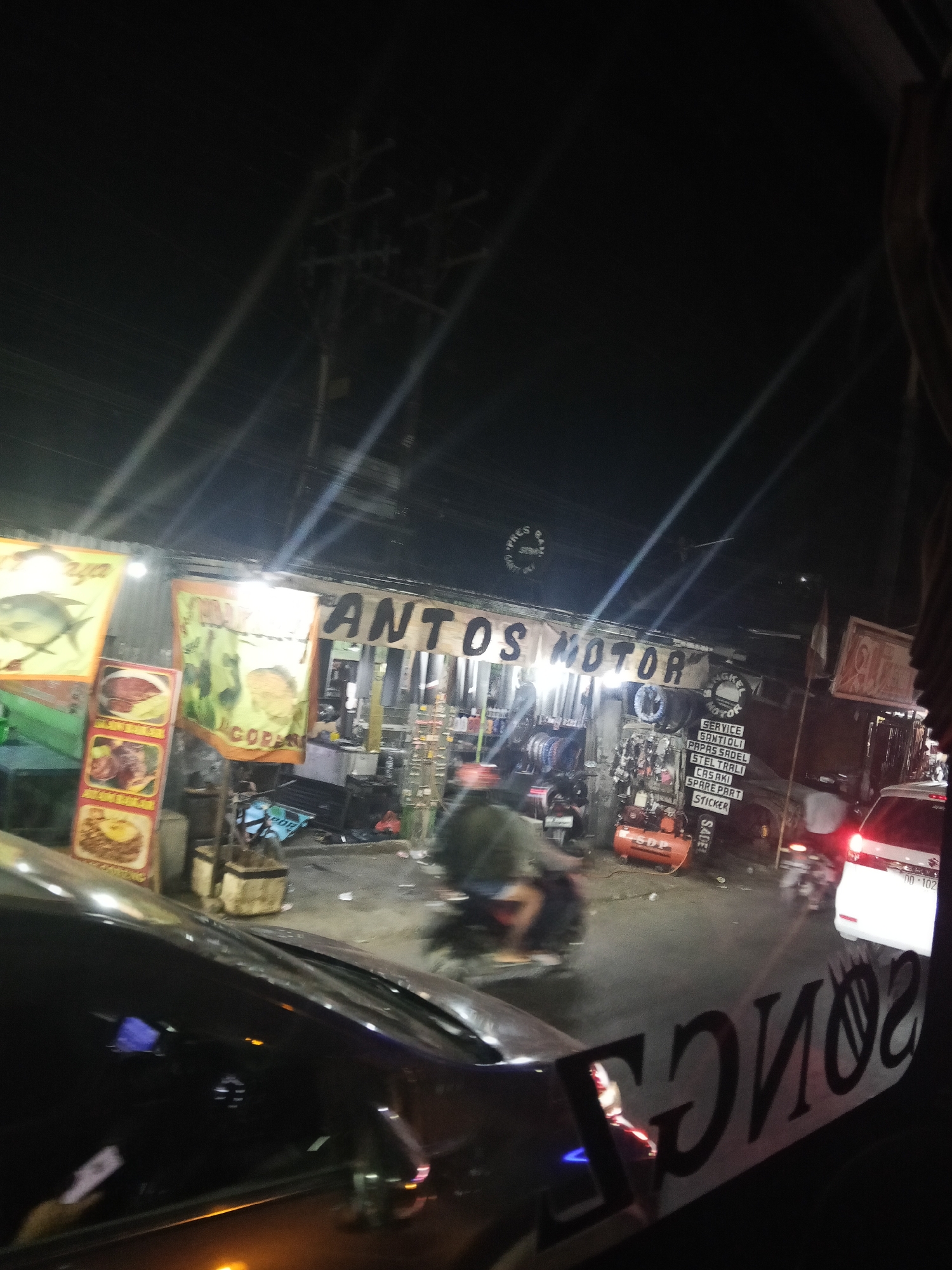 The image depicts a street scene at night in Marumpa, Indonesia, featuring a business named "ANTOS MOTOR." The storefront is illuminated, showcasing various motorcycle parts and accessories displayed on shelves and hanging from racks. A person is visible inside the establishment, possibly an employee. Outside, a motorcycle is being ridden by an individual wearing a green jacket and blue shorts. A white van is parked on the right side of the street, with its brake lights illuminated. The street itself appears wet, suggesting recent rain. A black car, possibly the perspective from which the photo was taken, is in the foreground, with a reflection of lettering on its window. The overall atmosphere is that of a busy commercial area at night.