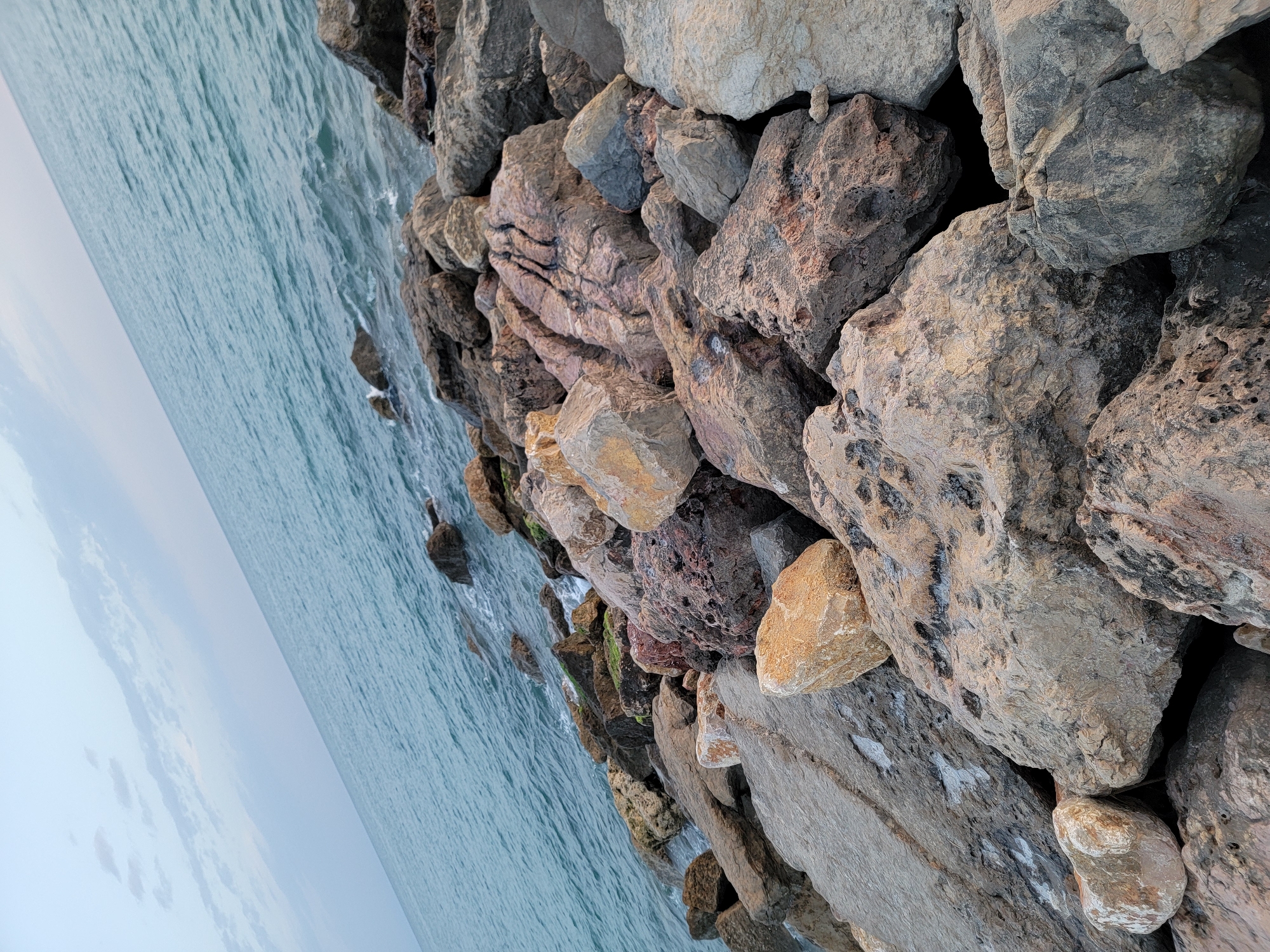 A rocky shoreline is depicted, with the ocean meeting the land. The image is taken from a low angle, looking up at the sky and across the water. The rocks are of various sizes and colors, ranging from light gray to dark brown and reddish-brown. Some of the rocks are covered in moss or algae. The ocean is a deep blue, with small waves lapping at the shore. The sky is a pale blue with scattered clouds. The overall impression is one of a natural and rugged landscape.
The location is the Quarteira shoreline in Portugal, where large rocks have been placed to form a barrier against the sea.