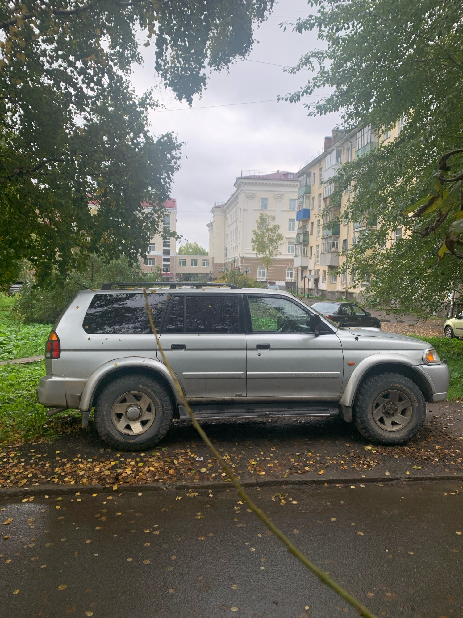 An overcast day in Arkhangelsk, Russia, captures a silver Mitsubishi Pajero parked on a street lined with apartment buildings and trees. Fallen yellow leaves are scattered across the wet asphalt and the curb, indicating an autumn season. The car, a sport utility vehicle, is positioned on the right side of the frame, facing left. Its windows are tinted, and its tires appear rugged, suggesting it might be used for off-road activities.
In the background, several apartment buildings are visible. The closest one is a light yellow, multi-story structure with balconies, some of which are enclosed with glass. To the left, another building with a reddish roof and decorative white columns stands out. Trees with green and yellow foliage frame the scene, some branches hanging over the car and street. A thin, branch-like object extends diagonally from the bottom right corner towards the center of the image, partially obscuring the car. The street is wet, indicating recent rain. No people are visible in the image.