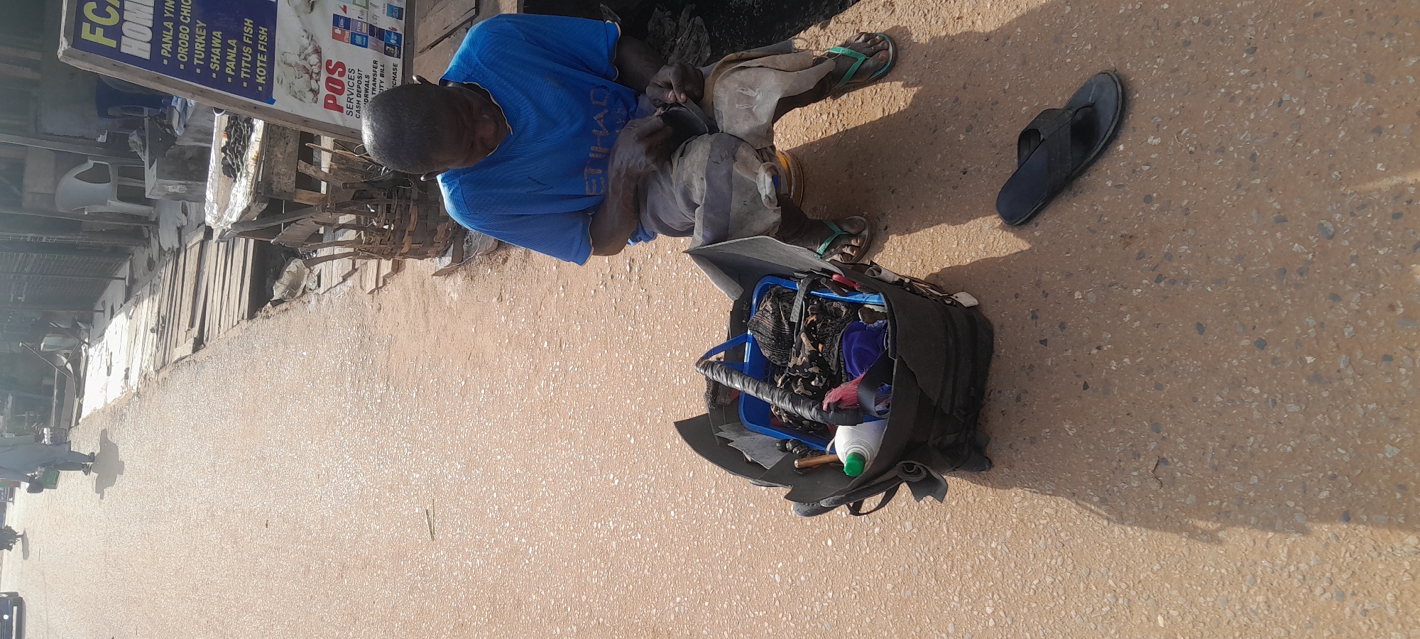 An outdoor, eye-level, medium shot captures an elderly African man in a blue t-shirt and grey trousers sitting on the ground in Abuja, Nigeria. He is actively engaged in repairing something with his hands, which are obscured by his clothing and the object he is working on. To his left, a large, dark-colored bag with various items protruding from it is situated on the ground. Among the visible items are a blue plastic container, what appears to be a bottle with a green cap, and various fabrics. A black flip-flop lies on the ground a short distance in front of the man. In the background, a wooden structure and a sign with text are visible, along with some fencing and a white chair. The ground is a mixture of asphalt and sand, with scattered debris. Sunlight casts shadows on the scene.