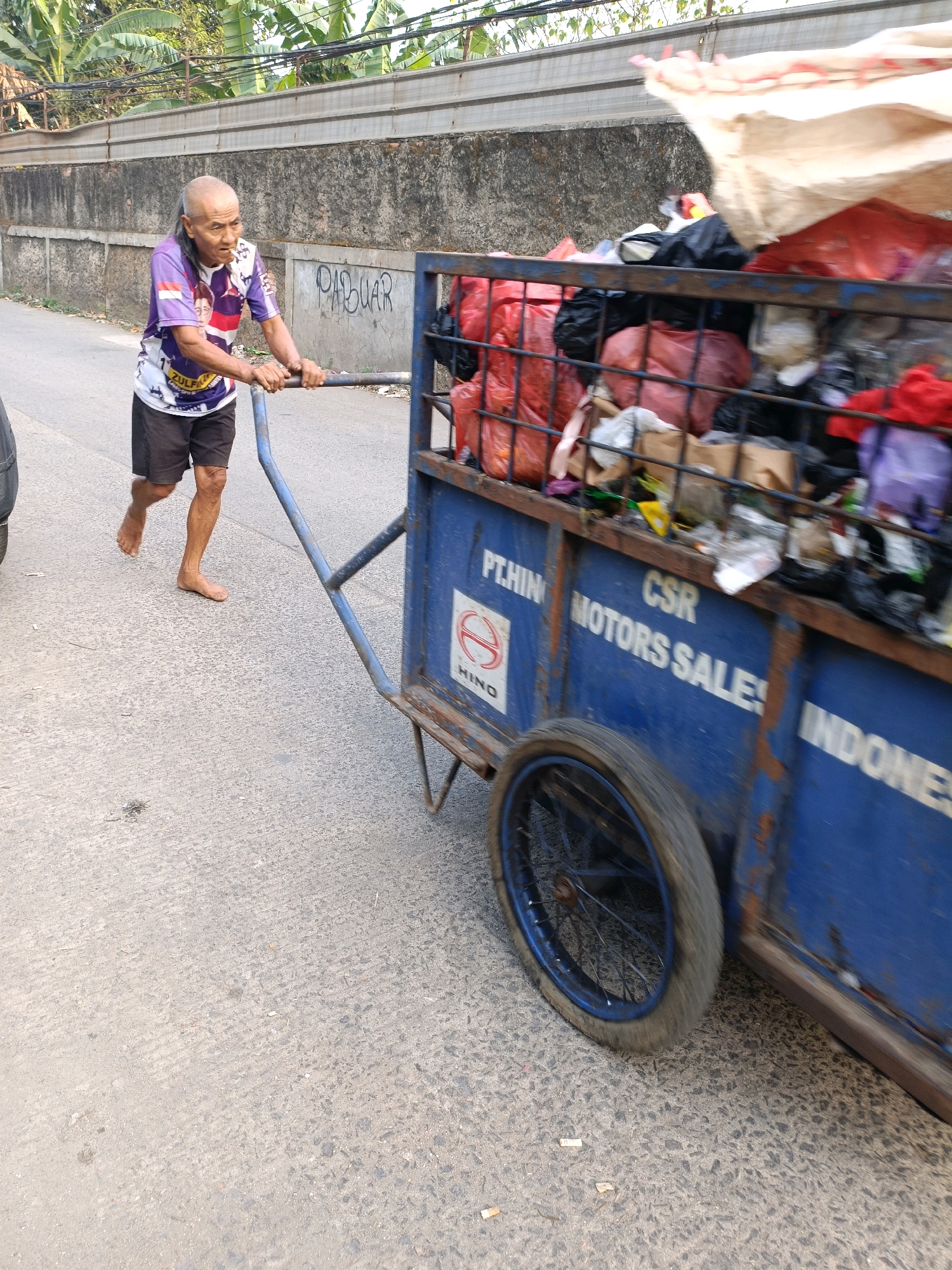 An elderly, bald man with a long ponytail is pushing a blue, two-wheeled cart filled with refuse. The man is wearing a purple t-shirt with images of faces and Indonesian flags on it, along with dark shorts. He is barefoot. The cart has "PT. HINO CSR MOTORS SALE! INDONESIA" written on its side, along with the Hino logo. The cart is laden with various items, including red and black plastic bags, some containing what appear to be clothing or textiles, and a large cream-colored sack on top. The scene takes place on a paved road with a concrete wall in the background, which has graffiti on it that reads "PASAR". Lush green vegetation is visible above the wall. The environment suggests a typical street scene in the city of Tangerang, Indonesia.