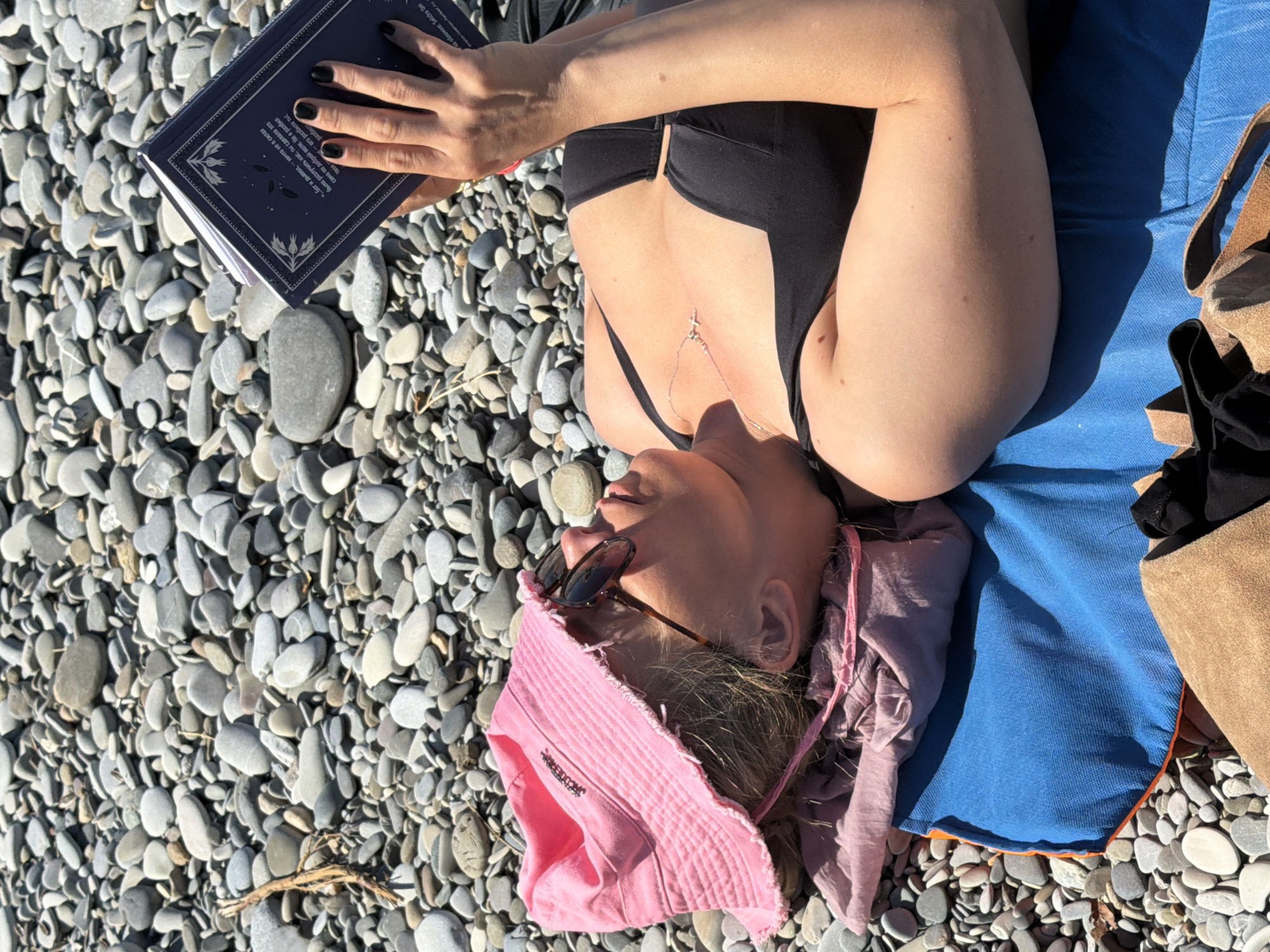 A woman lies on a blue towel and a pink cloth on a pebble beach. She is wearing a black bikini top, sunglasses, and a pink bucket hat. She is holding a dark blue book open in her right hand, and her nails are painted black. The beach is covered in grey and white pebbles, with some dry branches scattered around. The woman is looking at the book. The location is Mamedova Shchel', Russia.