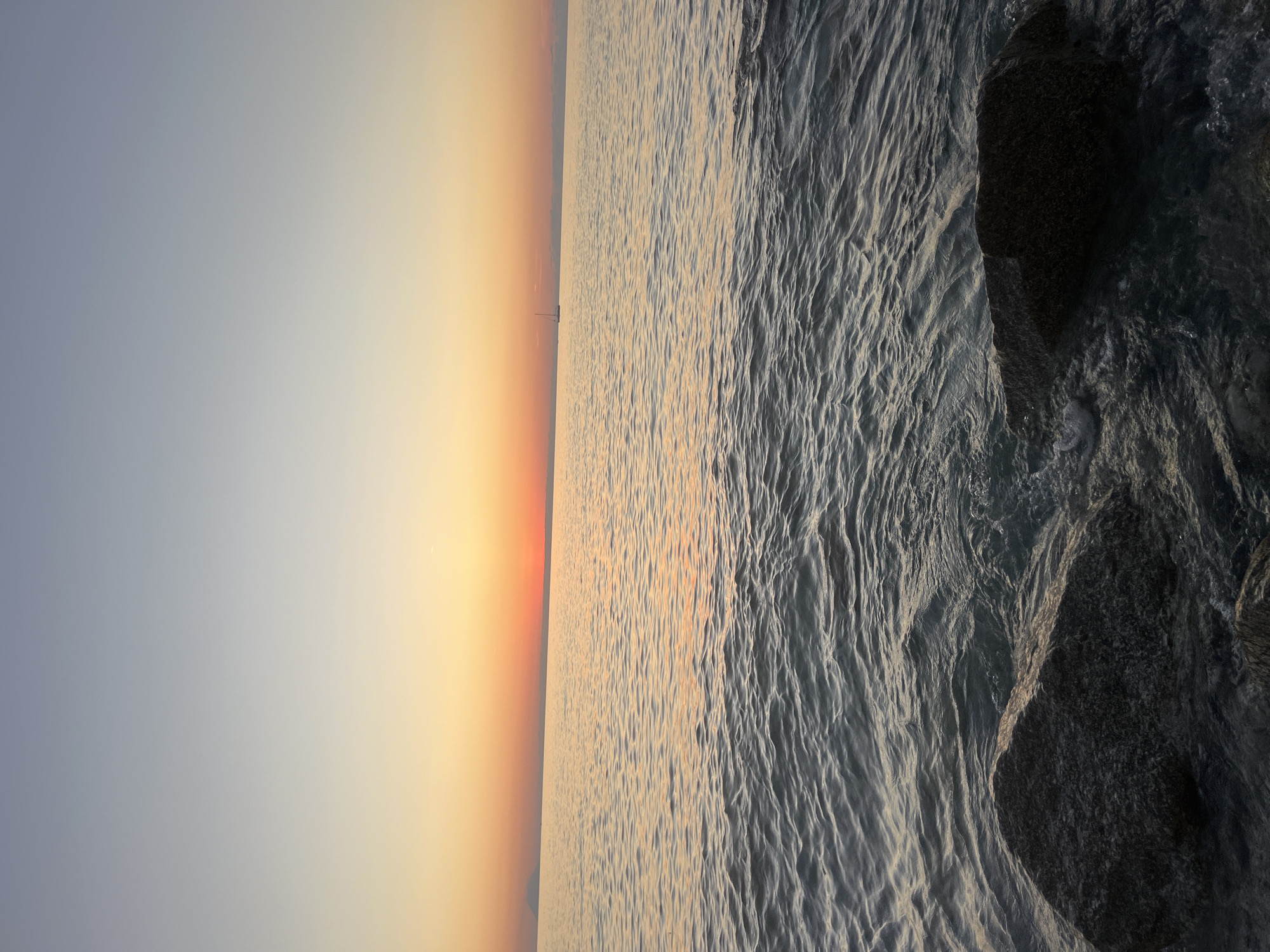 This image depicts a serene seascape at sunset, likely near Tropea, Italy, given the characteristic calm waters and clear skies of the Tyrrhenian Sea. In the foreground, dark, wet rocks are partially submerged in the gentle waves, their textures highlighted by the soft light. The water itself reflects the warm hues of the sunset, creating shimmering patterns on its surface. In the middle distance, a lone sailboat glides across the calm sea, its silhouette a subtle detail against the vibrant horizon. The sky transitions from a pale blue at the top of the frame to soft oranges and pinks closer to the horizon, where the sun has recently set. The overall impression is one of tranquility and natural beauty.