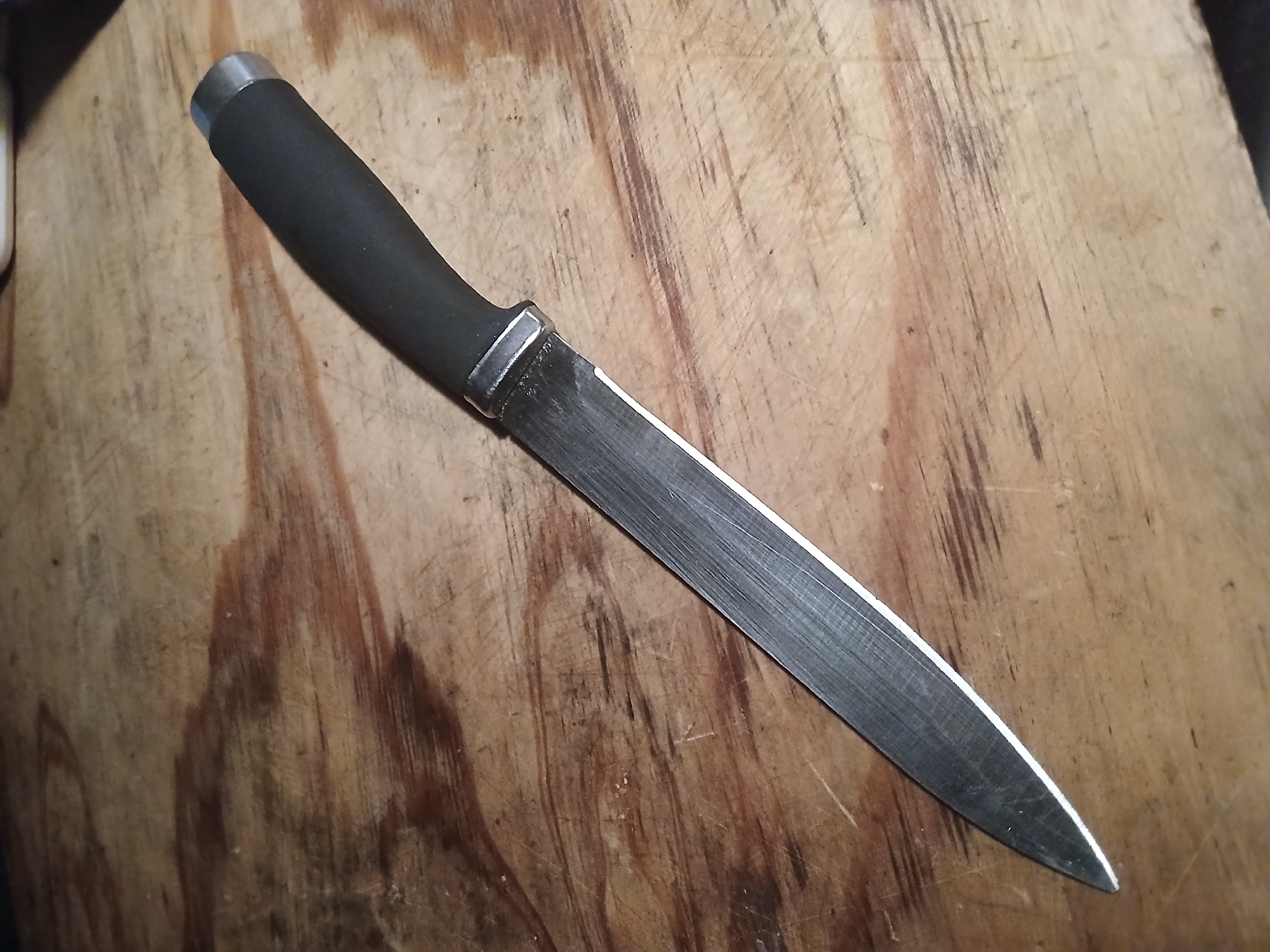 A knife with a black handle and a metal pommel lies diagonally on a weathered wooden surface. The knife's blade is long and narrow, with visible striations from sharpening. The wooden surface exhibits a grain pattern and appears to be a table or workbench. The lighting is somewhat dim, creating shadows that emphasize the texture of the wood. There are no people or other objects in the frame. The image was taken in Douai, France.