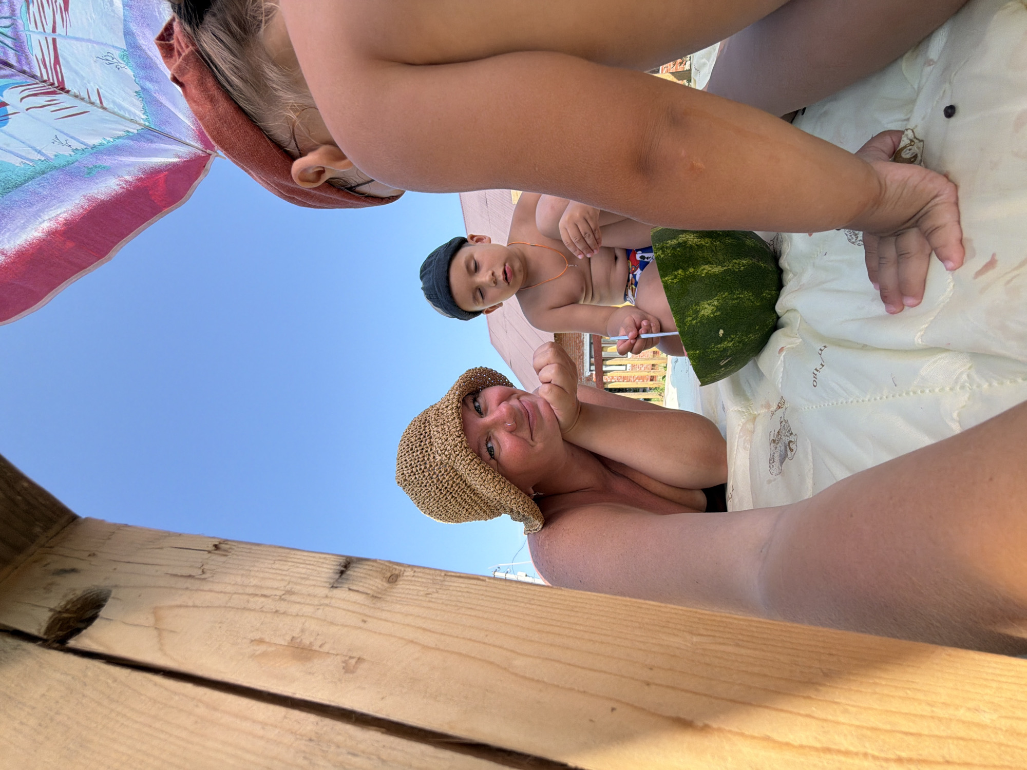 A low-angle, close-up shot captures a group of people enjoying a day outdoors, likely a beach or picnic setting, given the presence of a beach umbrella and a slice of watermelon. The scene is bathed in bright sunlight, suggesting it is daytime, with a clear blue sky visible.
The main subjects include at least two individuals, with another child partially visible. One person, wearing a straw hat, is in the foreground, looking towards the camera with a relaxed and happy expression. They are resting their head on their hands and appear to be smiling. Another person, a young child wearing a dark beanie and patterned swim trunks, is seated further back, appearing to be engrossed in something. A large slice of watermelon is prominently displayed between them. The upper portion of the image shows the underside of a colorful beach umbrella, adding to the outdoor ambiance.
The overall mood is one of leisure and enjoyment, likely a family outing or gathering. The bright lighting and clear sky contribute to a cheerful atmosphere. The wooden structure in the foreground suggests a picnic table or a beach structure. While no visible text is present in the image, the context provided places the location in Chaikovsky, Russia.