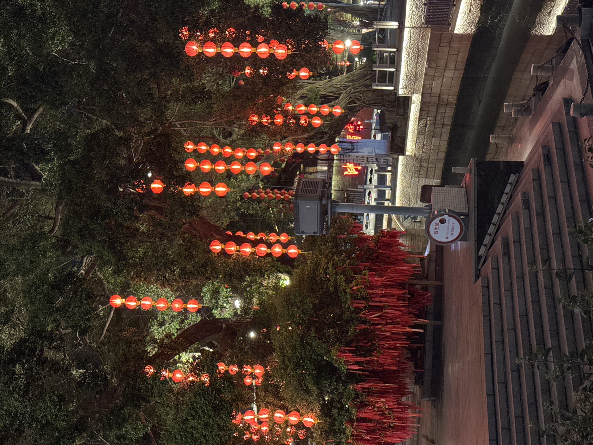 The image shows a nighttime view in Liwan District, China, featuring trees adorned with strings of red lanterns. The lanterns are evenly spaced, creating parallel lines of light. Adjacent to the trees is a brick wall covered in red ribbons or tags. A water feature or canal runs alongside the walkway, illuminated by lights. A sign with Chinese characters and the English word "LIFEBUOY" is visible on a post. The environment is dimly lit.