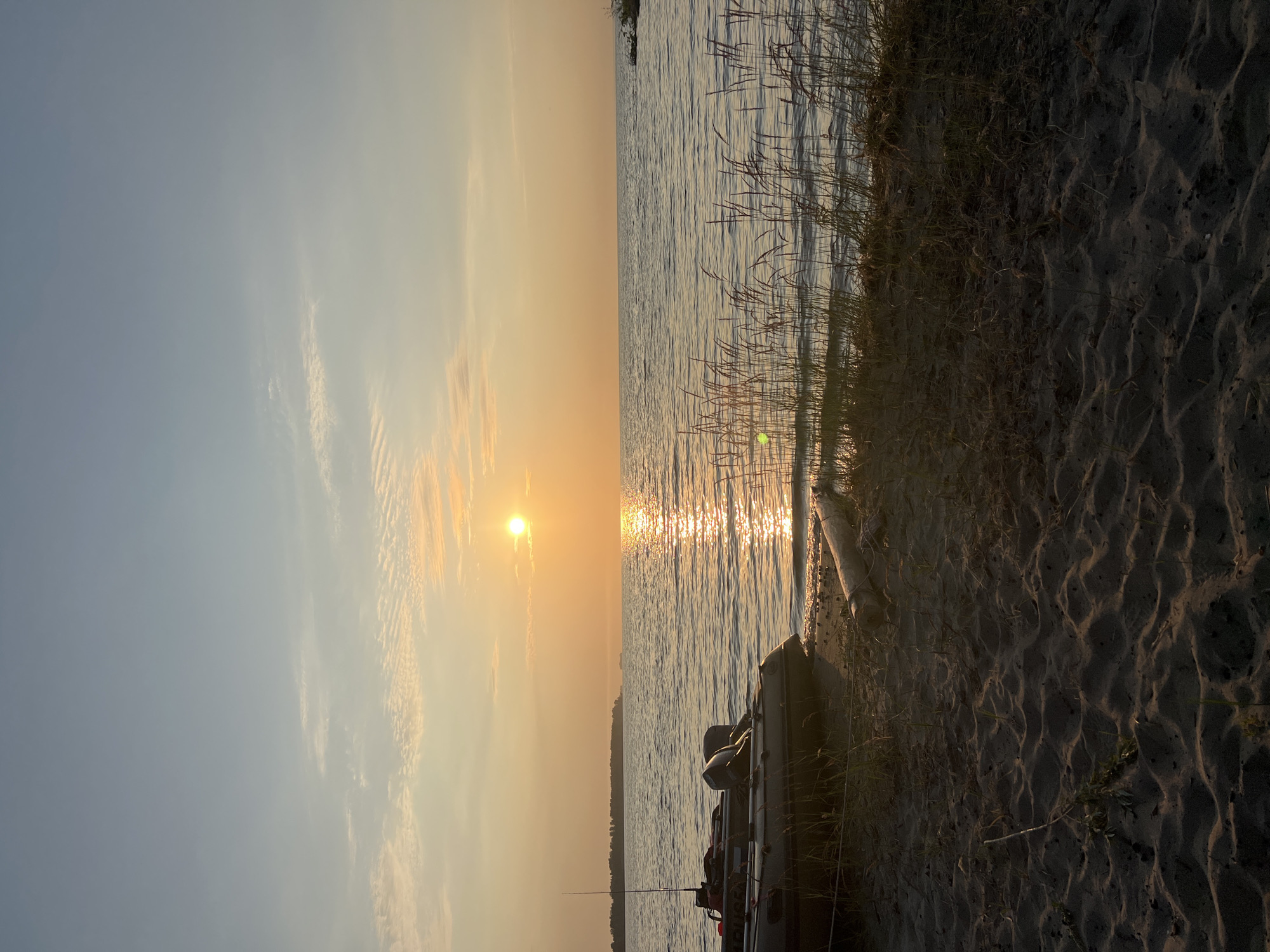 An inflatable boat with two outboard motors is parked on a sandy shore in front of a large body of water. The sun is setting over the water, casting a warm glow and creating a reflection on the surface. In the background, a tree-lined shoreline is visible, with a few distant structures. The sky is a pale blue with wispy clouds, and the overall mood of the image is peaceful and serene. The location is the city of Borovoe, Russia.