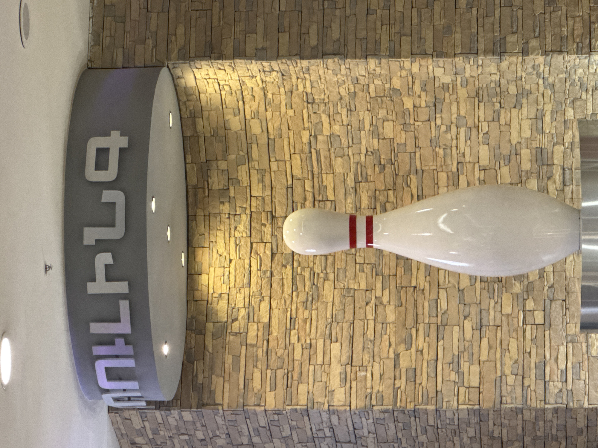 The image depicts a bowling alley in Yerevan, Armenia. A gray cylindrical sign with the word "Hollywood" in Armenian script is visible on the left. A large white bowling pin with two red stripes around the neck is positioned horizontally against a light brown brick wall. The environment suggests an interior setting, possibly a shopping mall or entertainment complex. There is no indication of people or actions in the scene.