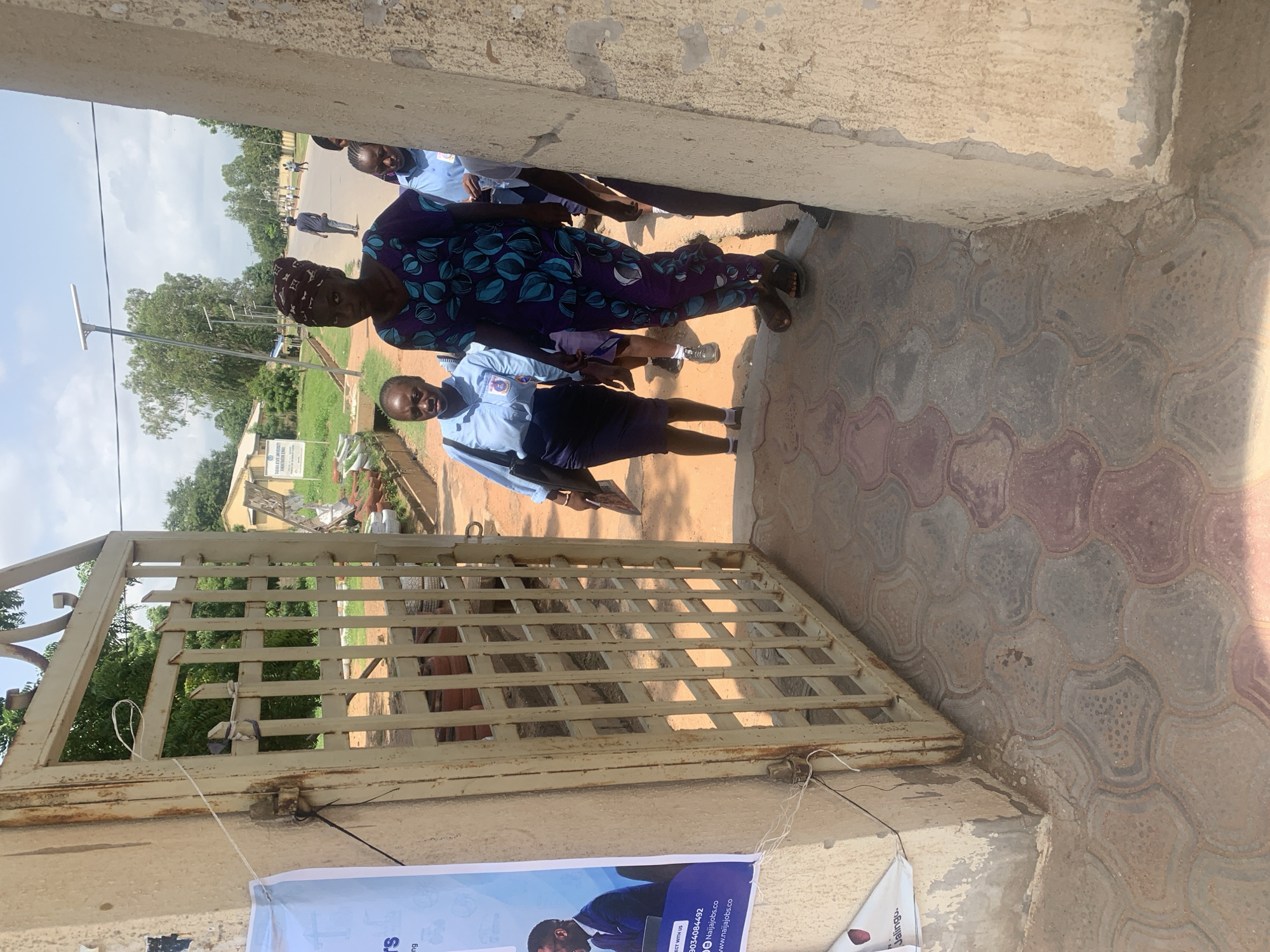 The image shows a group of people standing in front of a metal gate. There are at least five individuals visible, all appearing to be adults. One woman in the foreground wears a purple and blue patterned dress and a headscarf. She is looking towards the camera. To her left, a person in a light blue uniform with dark blue shorts is also looking at the camera. Another person in a similar uniform is visible behind them. Further in the background, more people in uniforms can be seen, some walking on a paved road. The scene appears to be outdoors, with trees and buildings in the distance, suggesting an institutional or public setting. The ground is paved with decorative interlocking stones. The time of day seems to be daytime, given the lighting conditions, and the weather appears to be clear. A banner is partially visible at the bottom of the image, with some text visible, including "NAIJJOBS.CO". The overall mood seems neutral or perhaps engaged, with some individuals looking directly at the camera.