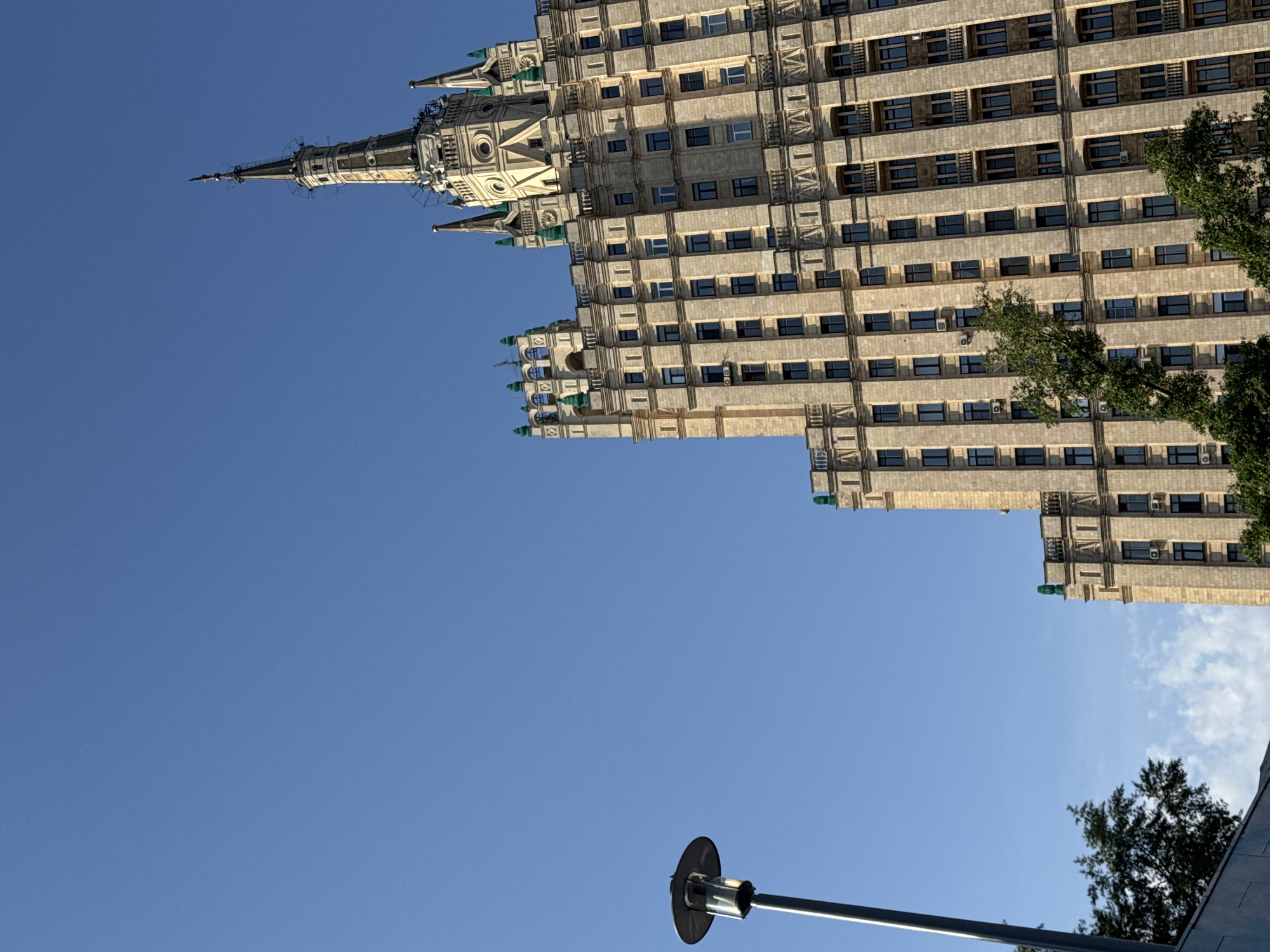 The image shows a view of the Hotel Ukraina, one of the Seven Sisters skyscrapers in Moscow, Russia. The building is characterized by its ornate Stalinist architecture, featuring a tiered structure, spire, and decorative elements. The facade consists of numerous windows. The photograph is taken from a low angle, emphasizing the height of the building against a clear blue sky. A lamppost is visible in the foreground. Trees can be seen adjacent to the hotel.