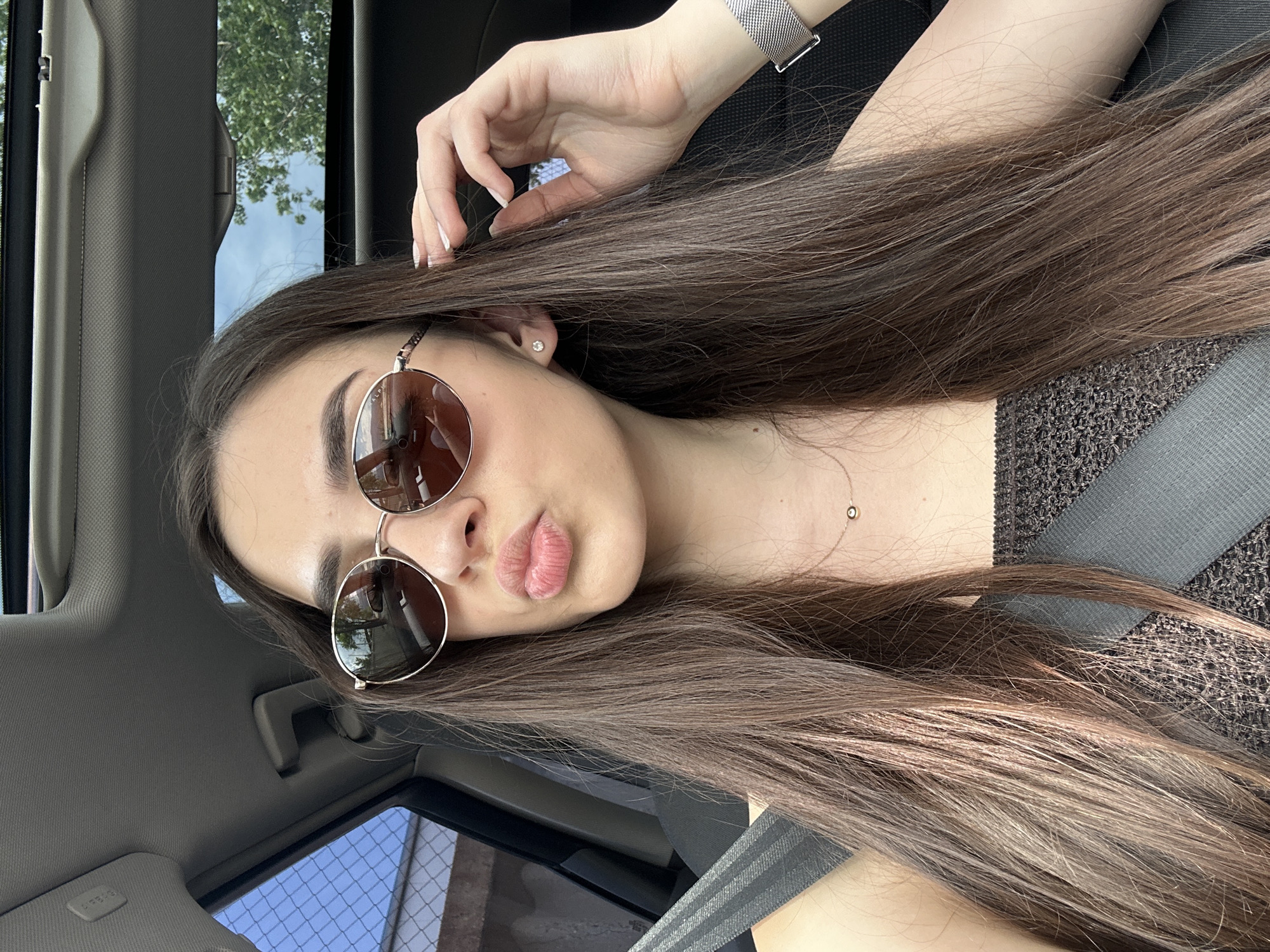 An individual is depicted seated in the front passenger seat of a vehicle in Unknown, Unknown. The subject is a female with long, straight, dark brown hair. She is wearing round, gold-framed sunglasses with dark lenses. Her lips are puckered in a "kiss" expression. A small, clear stud earring is visible in her left ear. She wears a delicate gold chain necklace with a small, dark circular pendant. Her attire includes a dark, possibly brown or black, textured or crocheted strapless top. A dark gray seatbelt is positioned diagonally across her chest and shoulder. Her left hand is raised, with fingers touching her hair near her left temple; her fingernails are painted a light color. A silver-toned mesh strap watch is visible on her left wrist. The car interior features a gray headliner and a visible sunroof through which blue sky and green tree foliage are discernible. Outside the car's side window, a chain-link fence and blue sky are visible.