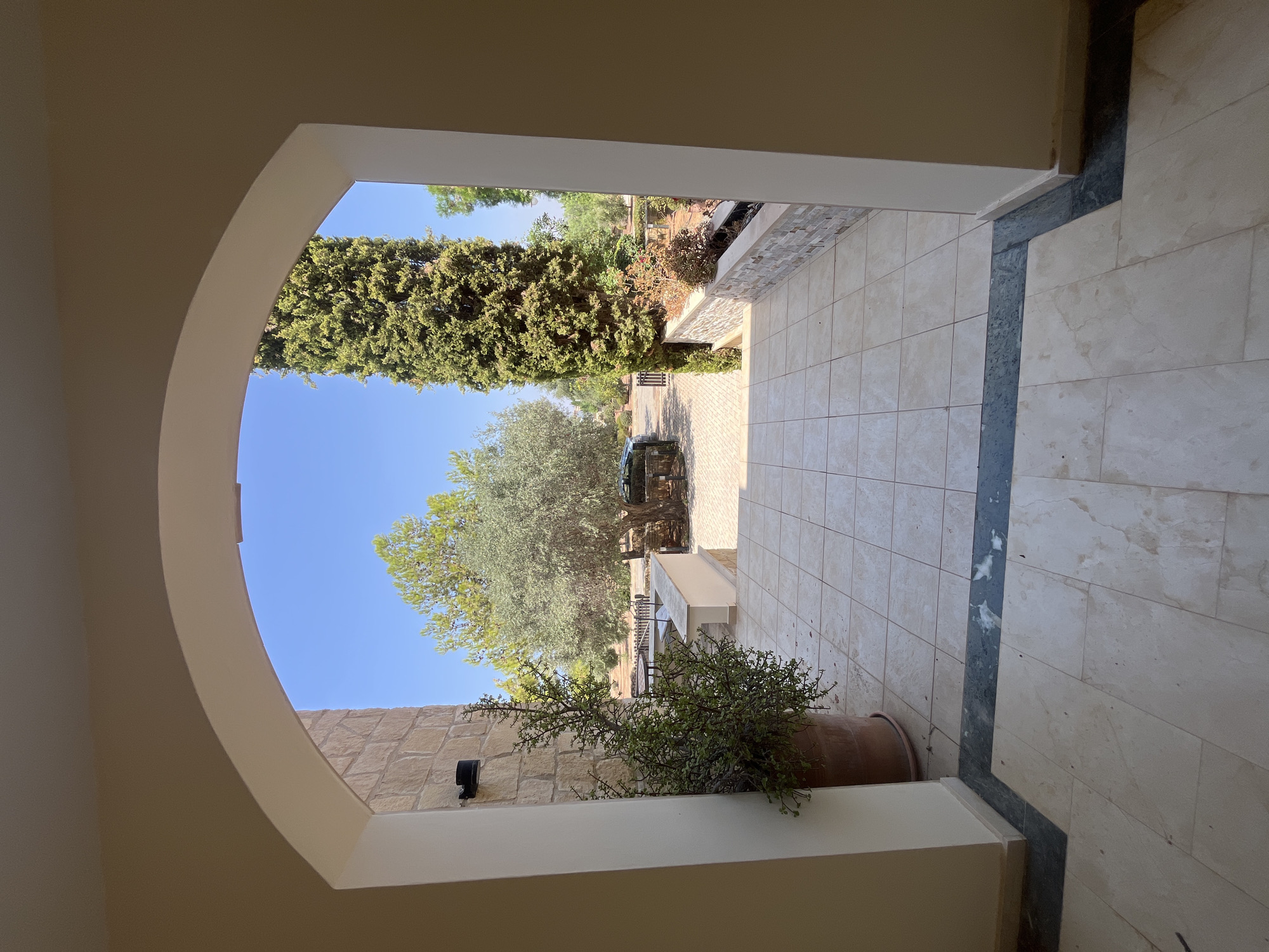 The image depicts an outdoor view framed by an archway. The foreground consists of a tiled patio with a decorative dark blue marble inlay. To the left, a large potted plant with green foliage stands near a stone-clad wall. Beyond the patio, a paved area leads to a green lawn with trees. A prominent tall cypress tree is visible on the right, adjacent to a low wall topped with stone and some dried plants. In the middle ground, an olive tree stands with a dark-colored car parked behind it. The background shows more greenery and a clear blue sky. The scene is consistent with a residential outdoor area, likely within a resort or private property, such as those found in Aphrodite Hills, Cyprus. No people are present.
