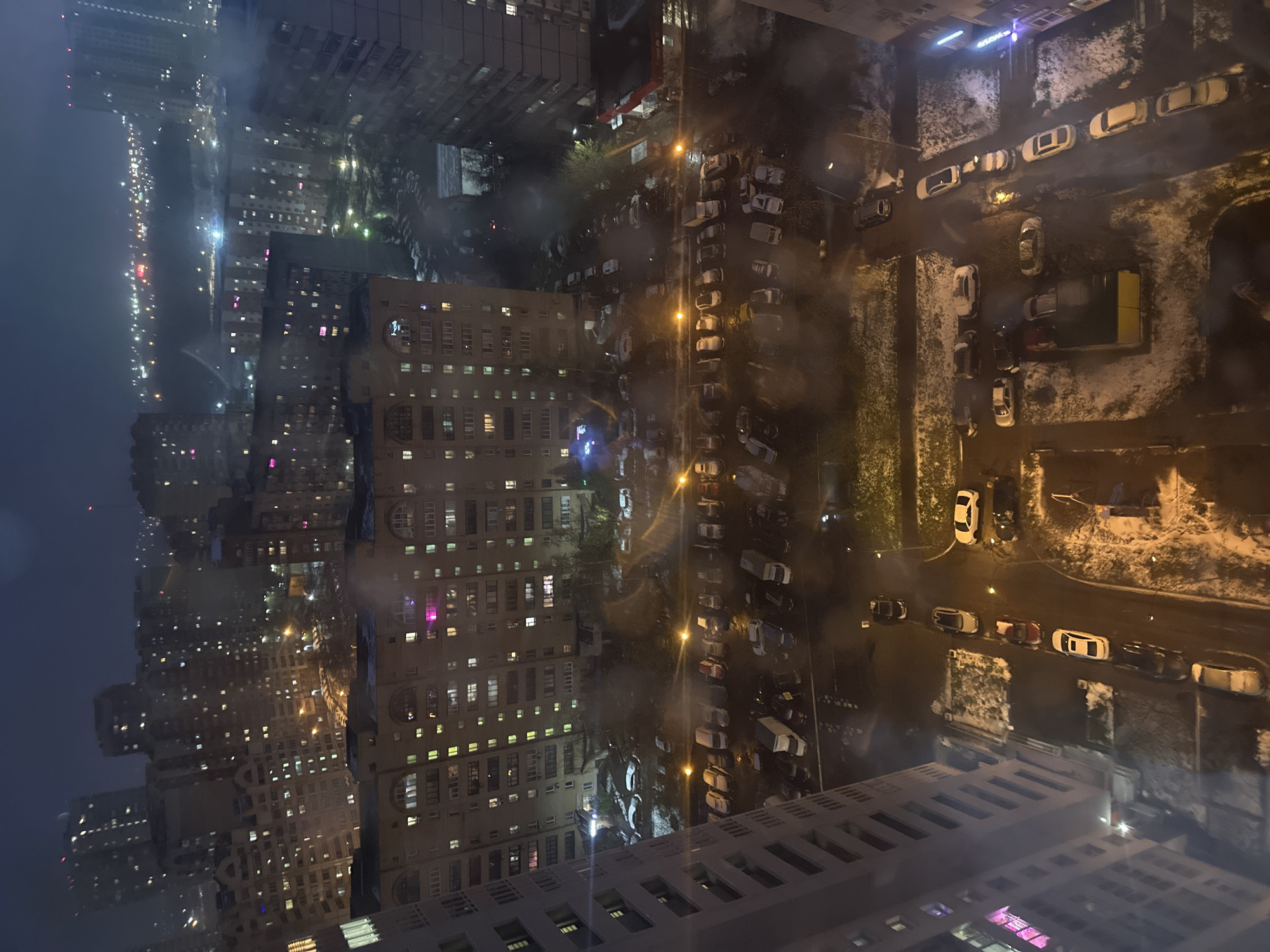 The media file displays a vertically oriented, elevated view of an urban residential area at night or early morning, likely captured from a high-rise building in Moscow, Russia. A significant amount of haze or fog permeates the scene, diffusing artificial lights and obscuring distant details, creating a soft, ethereal atmosphere.
Numerous multi-story apartment buildings dominate the landscape, their windows glowing with various lights – white, yellow, pink, and blue – indicating active residences. In the immediate foreground, the edge of another building, from which the photograph was taken, is partially visible. The architectural style of the buildings suggests a modern, dense city environment.
Below and between the buildings are roads and extensive parking lots, illuminated by streetlights that cast bright, warm glows. These areas are densely populated with parked cars, some covered with a light dusting of snow, confirming cold weather conditions. Patches of snow are also visible on parts of the ground, particularly in unpaved or less-trafficked areas. In the far distance, a dense cluster of city lights forms a bright, hazy line against the dark sky.
No people are discernible in the image, and no specific event or activity is taking place, presenting a quiet, nocturnal snapshot of urban life. There is no visible text in the frame.