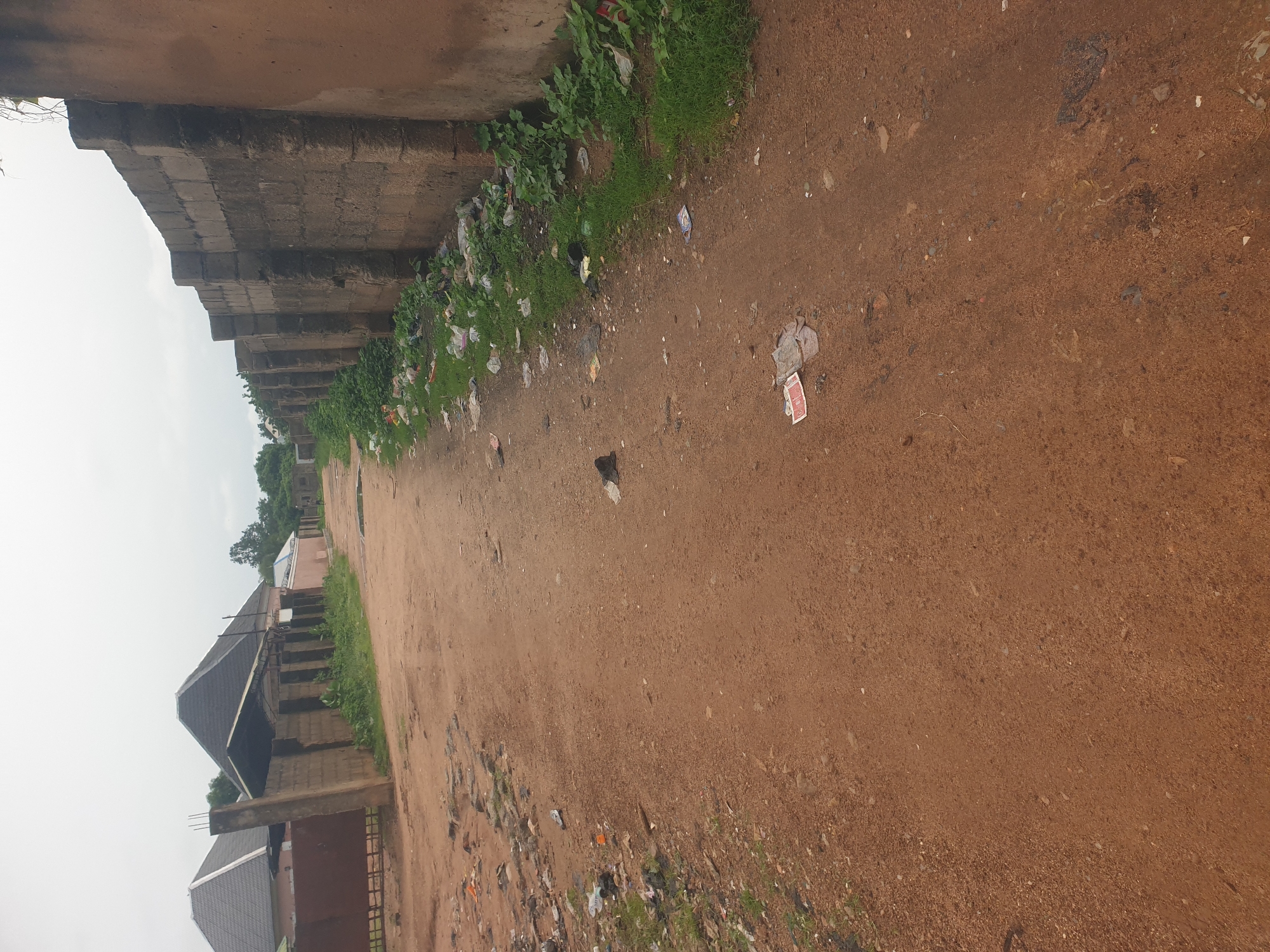 The image depicts an unpaved dirt road in a rural or developing area. The road is lined on one side by a tall, weathered brick wall that extends into the distance. On the other side of the road, there are several houses with pitched roofs, some appearing to be under construction or renovation. Fences and gates are visible in front of these properties. The roadside is littered with various debris, including plastic bags, paper, and other refuse, with patches of green grass growing amongst the trash. The sky is overcast.