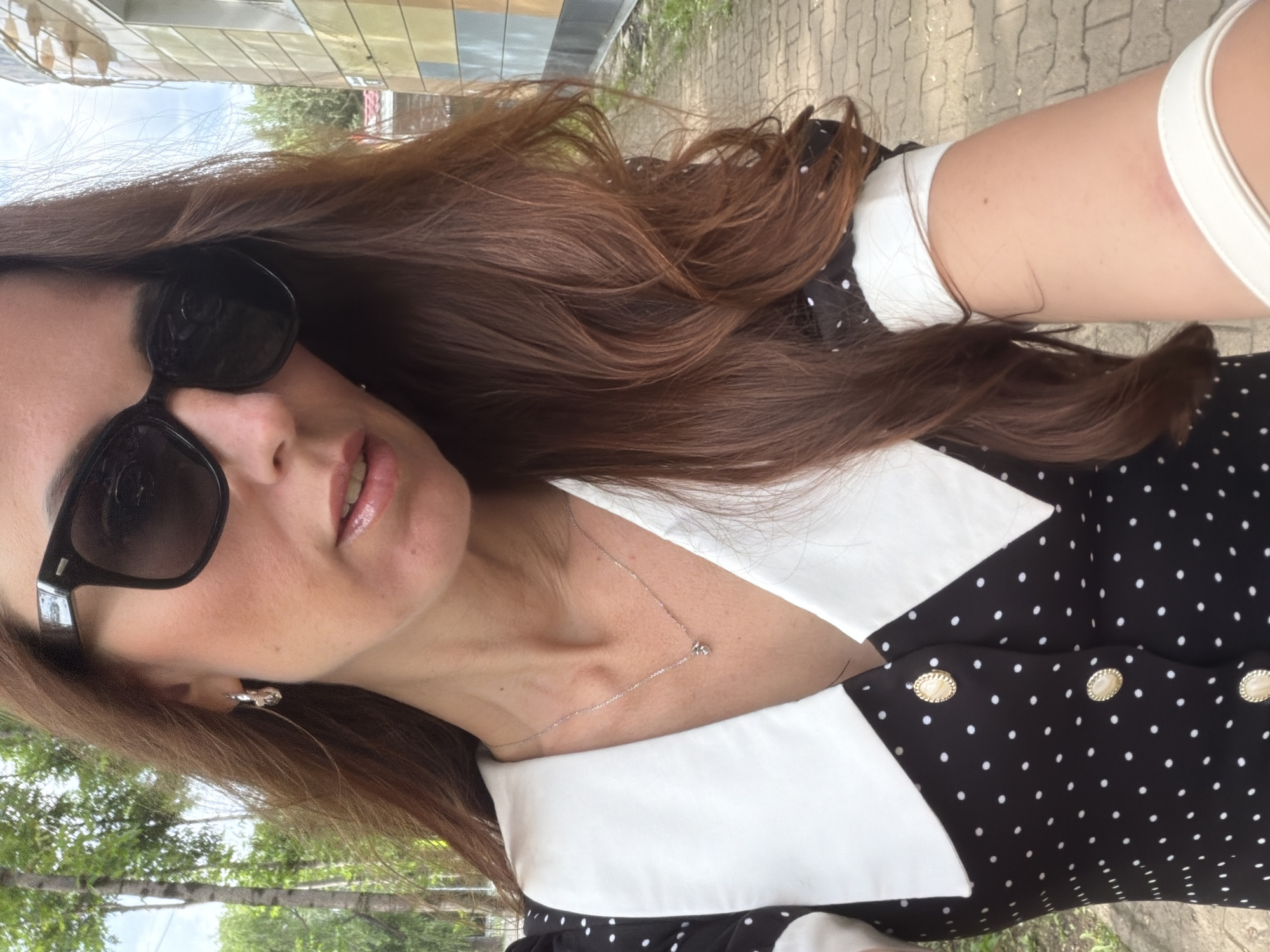 A woman with long, brown hair and dark sunglasses is posing for a selfie outdoors. She is wearing a black and white polka dot dress with a white collar and gold buttons. A delicate silver necklace with a small pendant is visible on her neck. The background shows trees and a building with a colorful facade, suggesting an urban or semi-urban environment. The cobblestone path and sidewalk indicate the location is in the city of Birobidzhan, Russia.