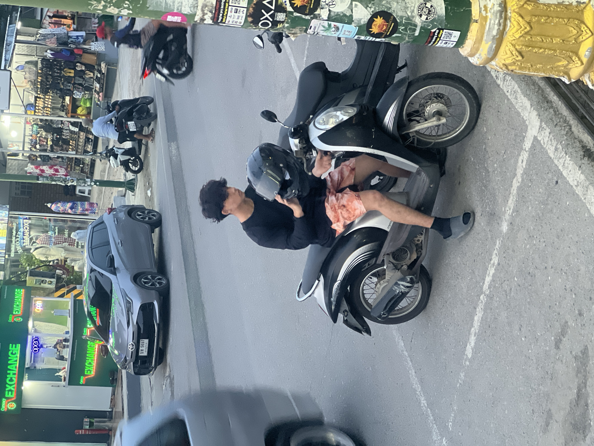 This image captures a street scene in the city of Ko Samui District, Thailand, likely near a commercial area with currency exchange services and shops. In the foreground, a young man with dark, curly hair is seated on a black and silver scooter, holding a black helmet in his hands. He is wearing a black long-sleeved shirt and light-colored shorts with a tie-dye pattern. The scooter has chrome accents and wire-spoke wheels.
To the left of the scooter, a silver car is parked, and a portion of a silver van or bus is visible, suggesting traffic. Behind the scooter, another scooter is parked, partially obscuring a black scooter with a person on it wearing a red cap. Further in the background, a building with neon green signage that reads "EXCHANGE" is prominent, indicating a financial service. Adjacent to the exchange, various shops display clothing and footwear. A lamppost with multiple stickers, including one for "TATTOO PIERCING," stands to the right of the image, framing the scene and providing context to the local environment. The ground is asphalt, marked with white lines indicating parking spaces. The lighting suggests it is daytime.