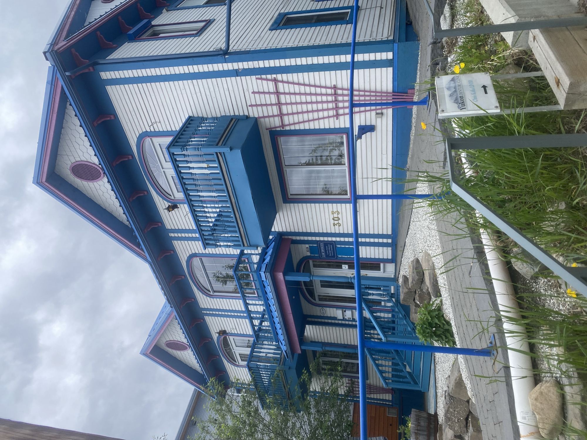 The image depicts the exterior of a two-story building in Whitehorse, Canada, with a distinctive white clapboard siding accented by bright blue and pink trim. The building features arched windows and two balconies with blue railings. A sign on the building reads "Law Offices" and "303." Another sign at the bottom right of the image says "Yukon Denture Clinic" and points with an arrow. The foreground shows a paved pathway and landscaping with rocks, grass, and some yellow flowers. The sky is overcast.
