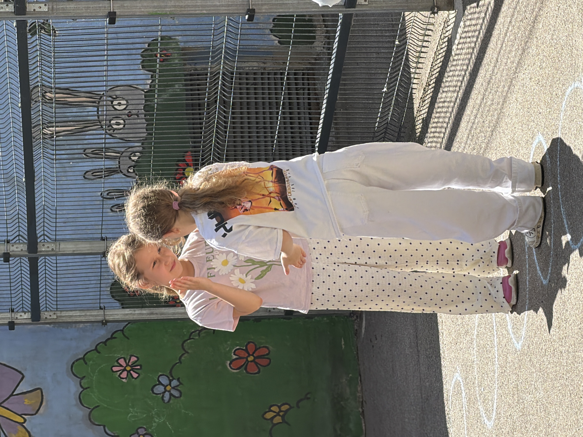 Two young girls are standing in an outdoor play area, facing each other. The girl on the left, with blonde hair pulled back, is wearing a pink t-shirt with a daisy design and white pants with black polka dots. She is holding her hand up near her face and looking at the other girl. The girl on the right, also with blonde hair in a ponytail, is wearing a white t-shirt with a colorful graphic and white pants. She is looking at the first girl and has her left hand extended towards her. Behind the girls is a metal fence. On the wall behind the fence, there is a mural depicting cartoon rabbits and greenery. The ground is asphalt with white chalk drawings of circles and lines, suggestive of a hopscotch or game area. Sunlight is casting shadows on the ground. The location is identified as being in the city of Riga, Latvia.