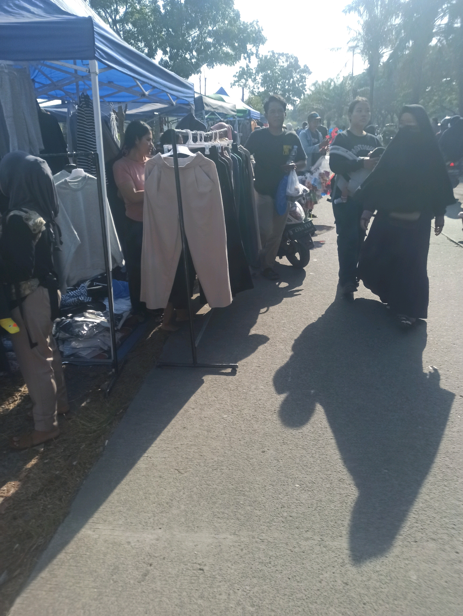 The image depicts an outdoor market scene in Telagamurni, Indonesia, characterized by a bright, sunny day and the presence of several individuals browsing or working at stalls. The foreground shows a paved road with tire tracks, and to the left, a grass verge bordering a clothing stall. The stall features a blue tarp canopy supported by poles, displaying various garments like t-shirts and pants hung on racks.
Several people are visible in the midground. On the left, a person in a dark headscarf and light-colored pants stands near the stall, appearing to be a customer. Next to them, a woman in a pink shirt looks towards the clothing. Further to the right, two men in dark t-shirts walk along the road, one carrying a plastic bag. Behind them, another person in dark clothing and a headscarf is seen, and a motorcycle is parked on the road. In the background, more stalls and trees are discernible, suggesting a bustling market environment. Shadows cast by the sun and the people indicate the time of day, likely late morning or early afternoon. The overall atmosphere appears casual and busy, typical of a local market.