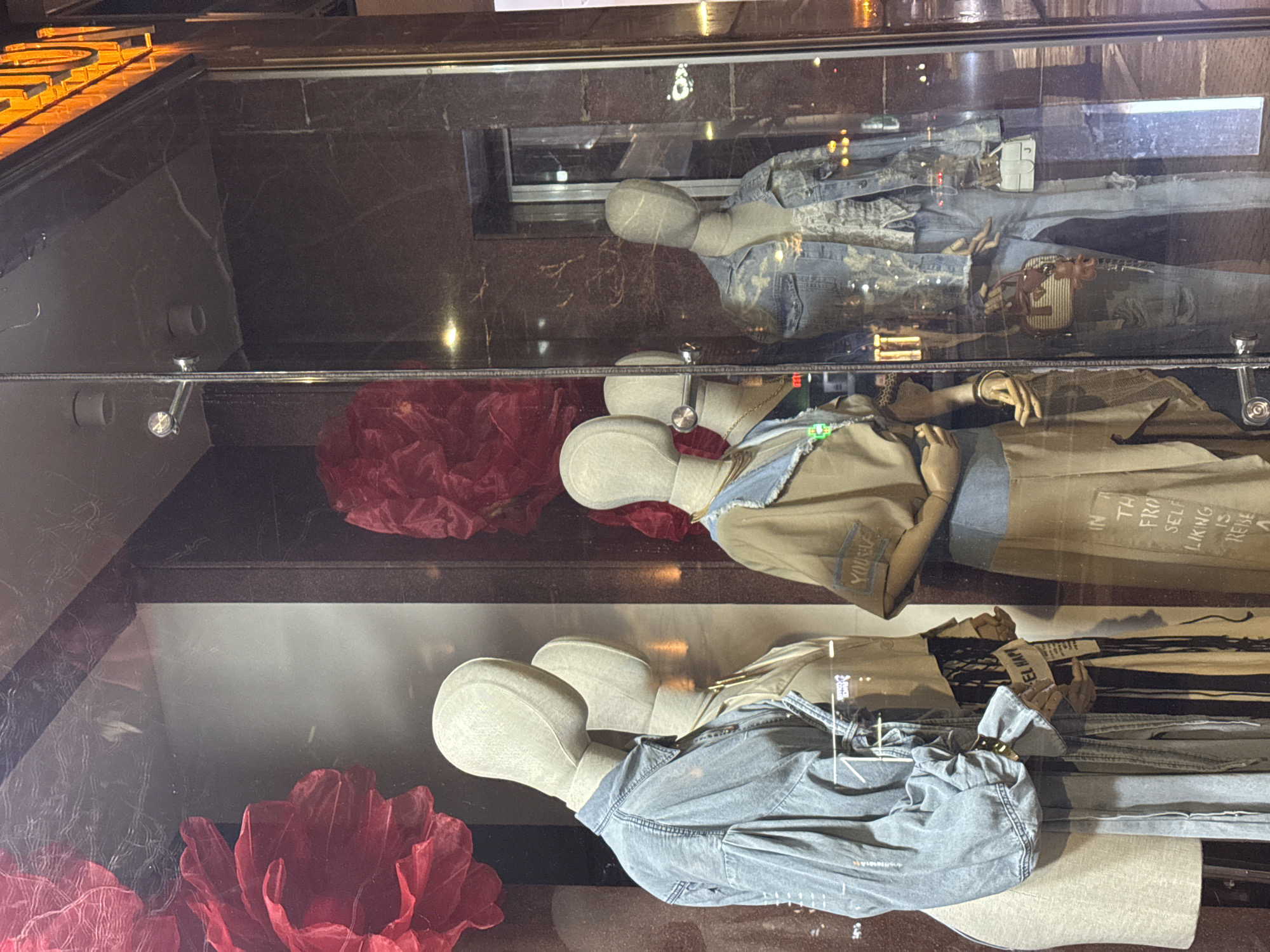 A window display in Yerevan, Armenia, features three headless mannequins arranged horizontally. The topmost mannequin wears a denim jacket and trousers. The middle mannequin is clad in a khaki jacket and trousers, featuring phrases like "IN THE FROM SELF LIKING IS REISE" printed on the garment. The bottom mannequin is dressed in a denim jacket. Large artificial red roses decorate the bottom left and middle left of the display. The reflection in the window shows city lights and surroundings.