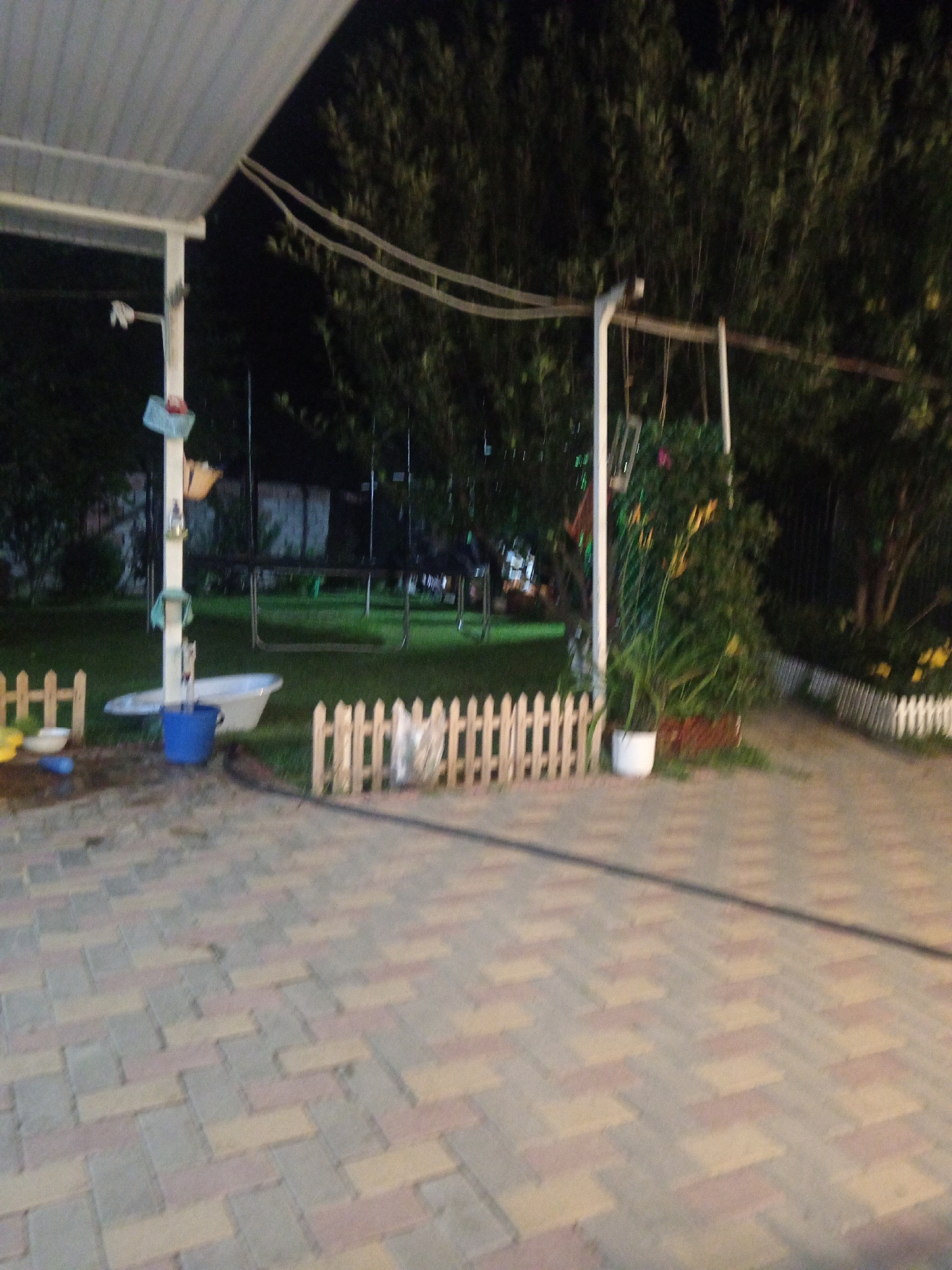 The image depicts an outdoor scene at dusk, possibly a backyard or garden area, illuminated by artificial lighting. A paved pathway made of rectangular stones in shades of gray, pink, and yellow is visible in the foreground, extending towards the right side of the frame. To the left, a white canopy structure with visible beams extends over the paved area. Adjacent to this structure, a white support post is adorned with several fabric items, including what appears to be a light blue towel and a darker blue bag. A white bathtub is positioned on the grass near the post, with a blue bucket placed next to it.
A low white picket fence runs horizontally across the middle ground of the image, separating the paved area from a grassy lawn. Beyond the fence, a trampoline with a metal frame is visible in the background, surrounded by trees and foliage that are dark with low light. To the right of the trampoline, a white vertical post supports a swing set, with chains and seats hanging down. In front of this swing set, a large white planter holds lush green plants, and further to the right, another section of white picket fence lines a path. The overall environment appears to be a residential outdoor space, possibly a suburban backyard or a communal garden area.