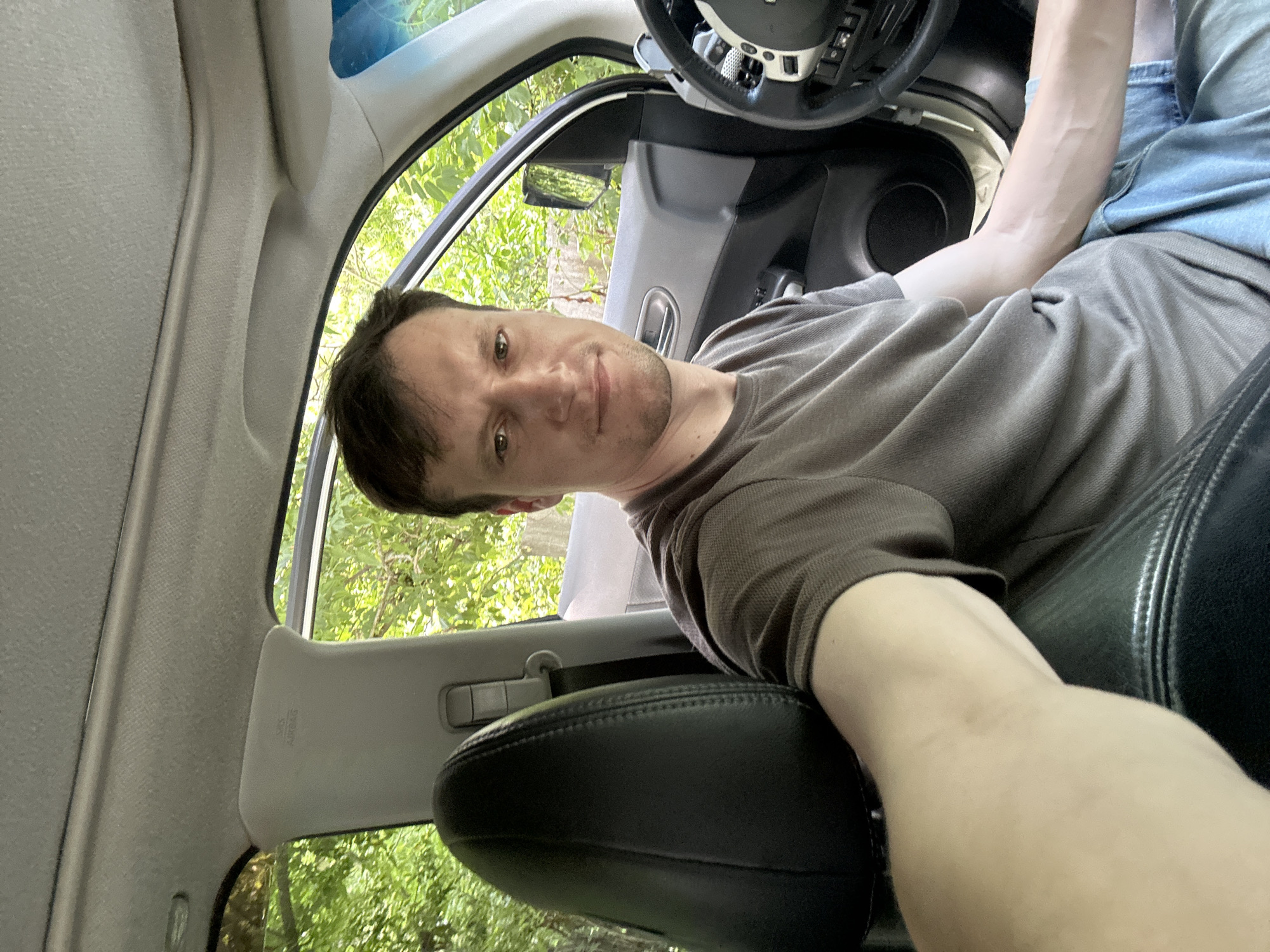 A man is taking a selfie in the driver's seat of a car. He is wearing a brown t-shirt and blue jeans. The car appears to be parked on a dirt road surrounded by trees. The interior of the car is visible, including the dashboard and steering wheel. The man has short brown hair and a light complexion. The image is taken from a slightly high angle, looking down at the man.