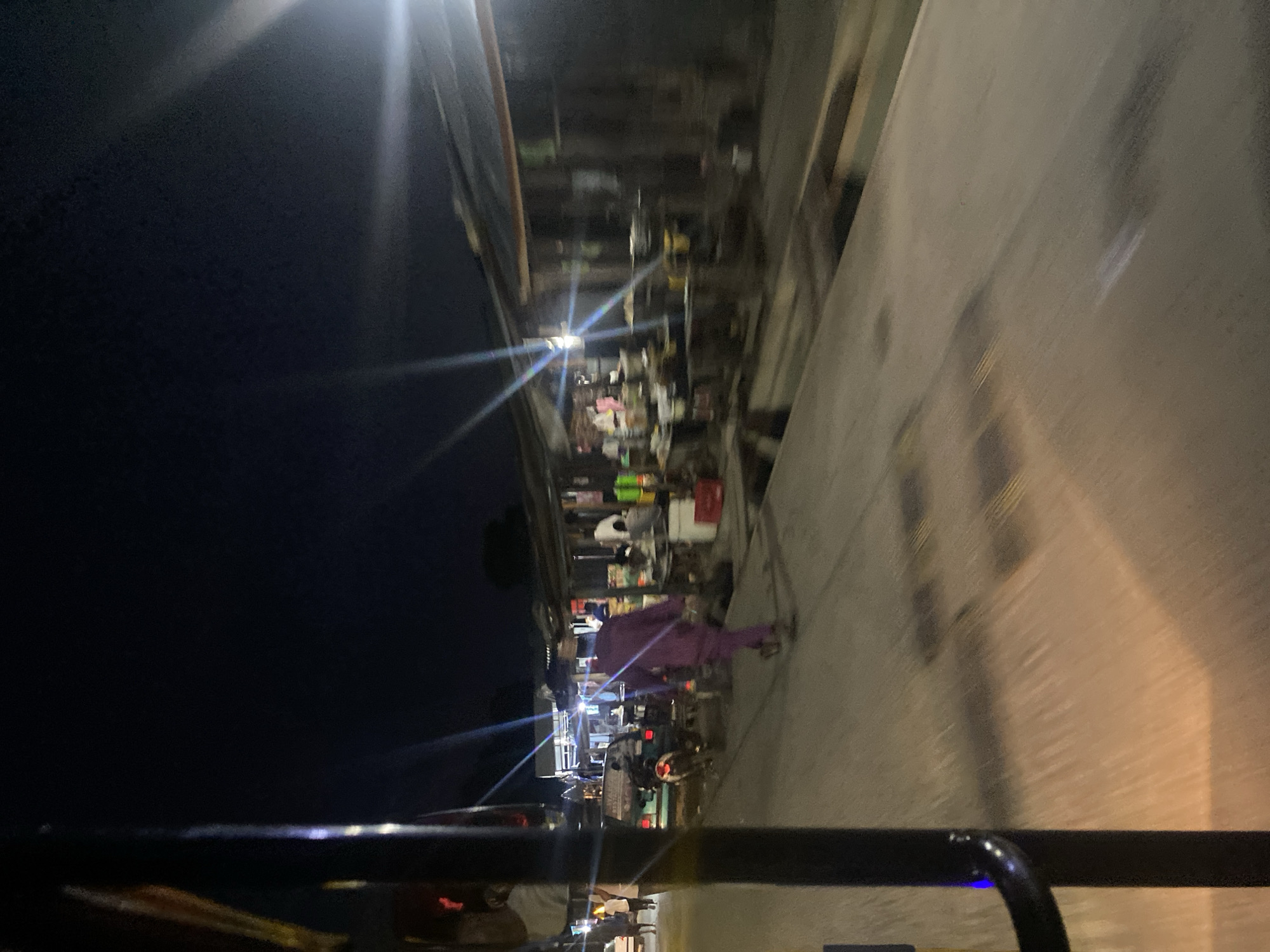 The image depicts a nighttime scene on a street in Jalingo, Nigeria. The perspective is from within a vehicle, looking out at a bustling roadside market. The scene is illuminated by artificial lights, including harsh streetlights creating lens flares and the glow from various stalls. The market appears to be selling a variety of goods, with shelves stocked with products and items displayed on tables.
Several people are visible. A person in a vibrant purple outfit is the most prominent human figure, walking or standing near one of the stalls. Other individuals are less distinct due to the low light and motion blur, suggesting activity and movement within the market. The presence of what appears to be a three-wheeled vehicle (an auto-rickshaw or similar) hints at local transportation.
The overall setting is that of a busy urban street at night, likely a commercial area where vendors set up shop after dark. The ground is paved, and a concrete structure, possibly a building or wall, runs along the side of the road. The quality of the image suggests it was taken with a handheld camera in low-light conditions, resulting in motion blur and some graininess. No specific weather conditions are discernible. No visible text is present in the image.
